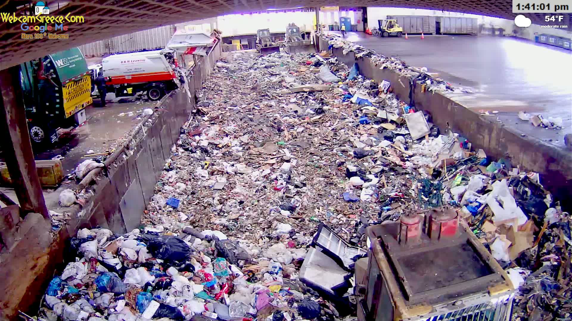A large pile of garbage sits in a waste disposal area under an overcast sky during moderate rain.