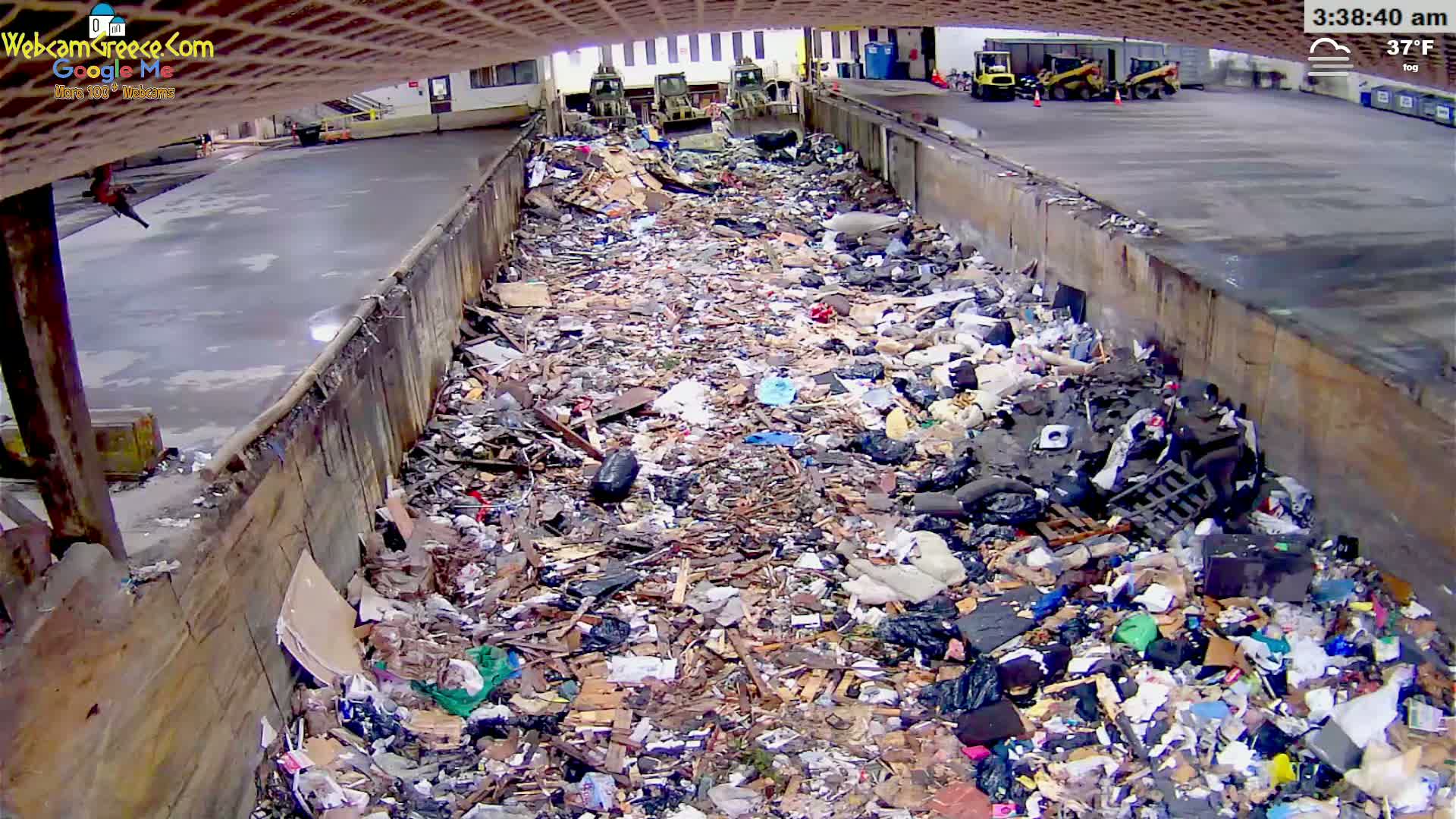 A large pile of trash fills a concrete trough outdoors in foggy, 37°F weather.