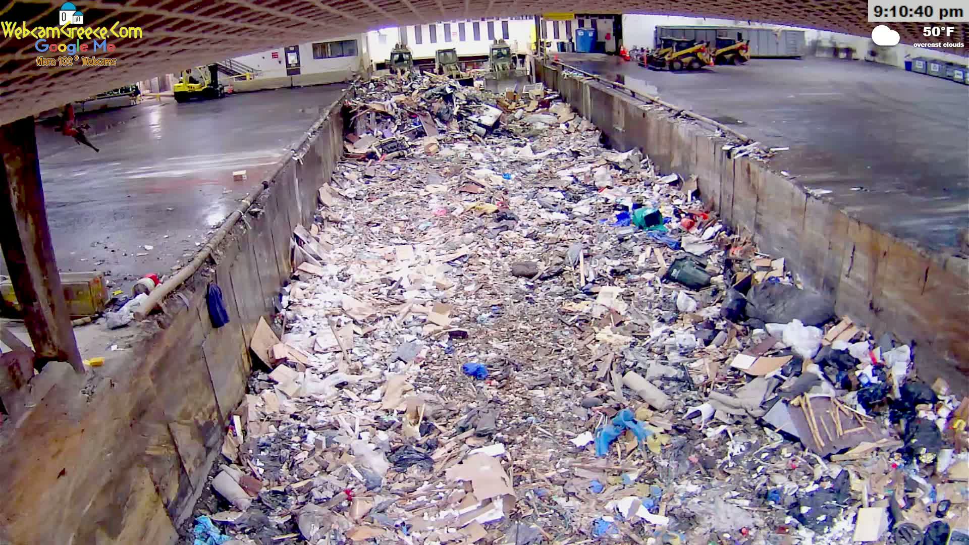 A large pile of refuse is seen in an outdoor concrete-walled bin under overcast skies, with several pieces of heavy machinery visible in the background.