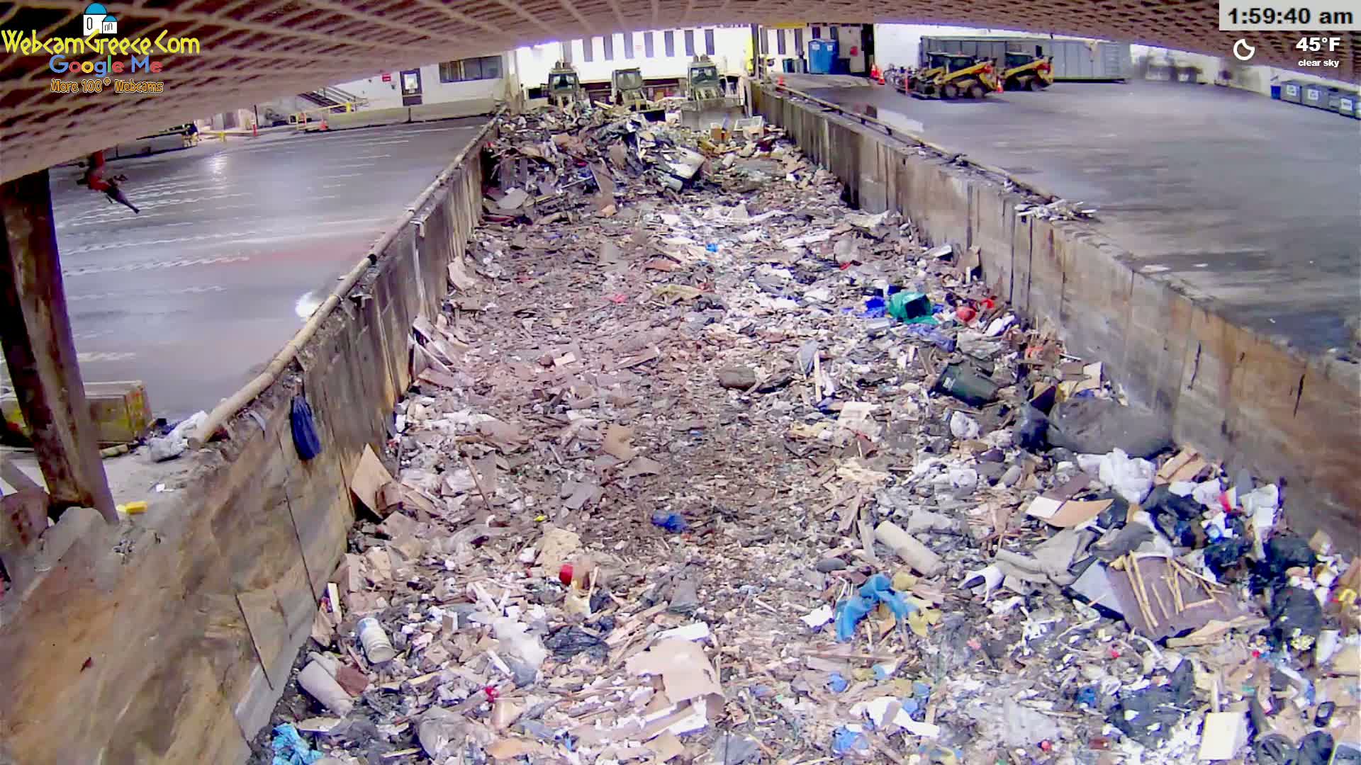 A large pile of debris and trash fills a long, narrow, walled-in area outdoors under a clear sky at 45°F.