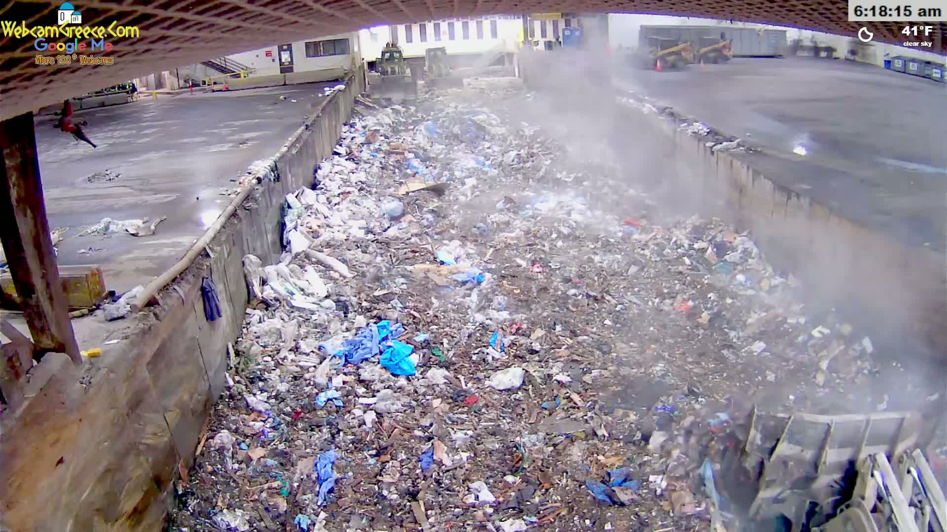 A large pile of trash and debris is being compacted by machinery in a waste processing facility on a clear, 41°F morning.