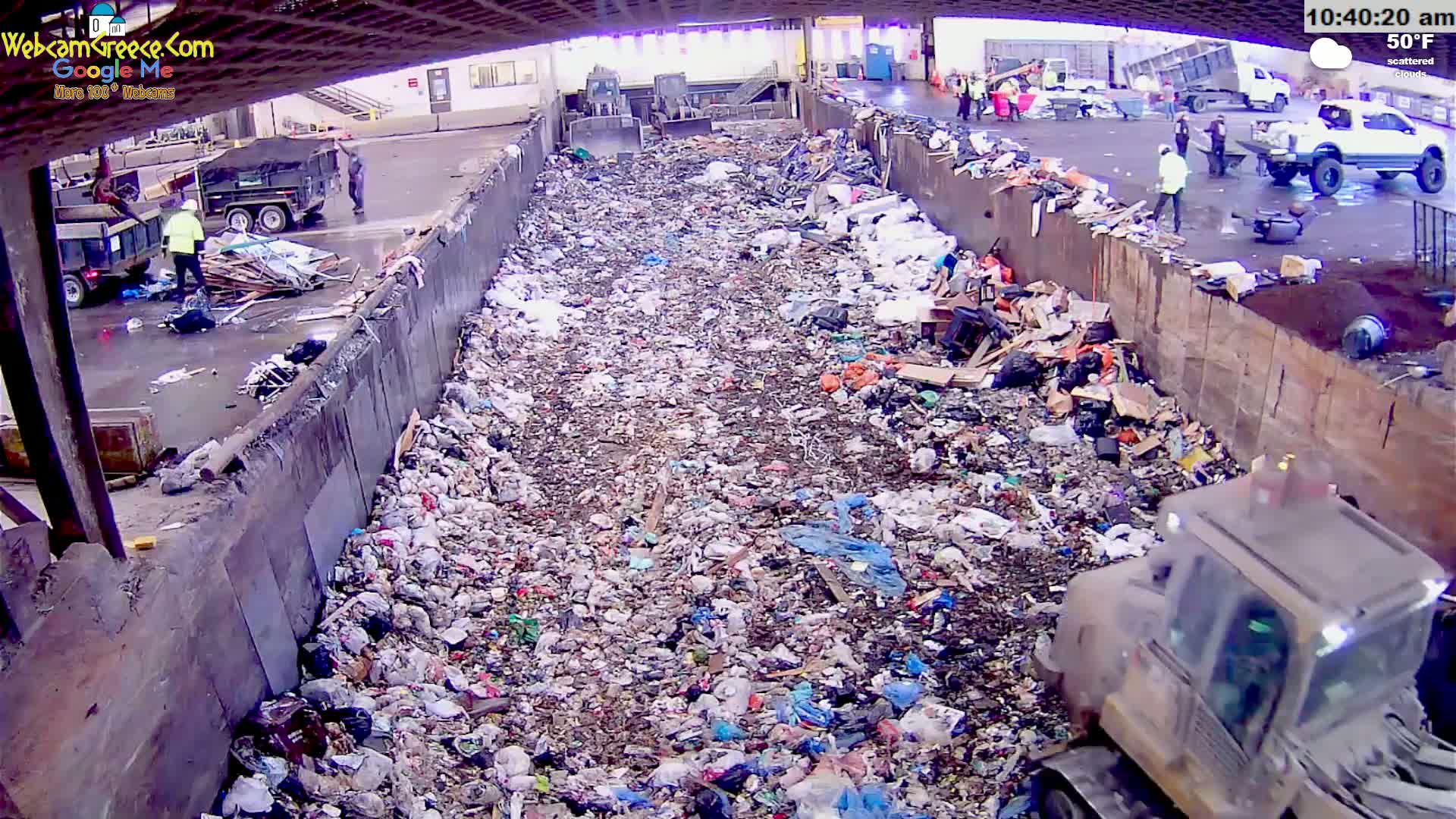 A large amount of trash is piled in a concrete-walled area under scattered clouds and a 50°F temperature, with workers and machinery visible nearby.