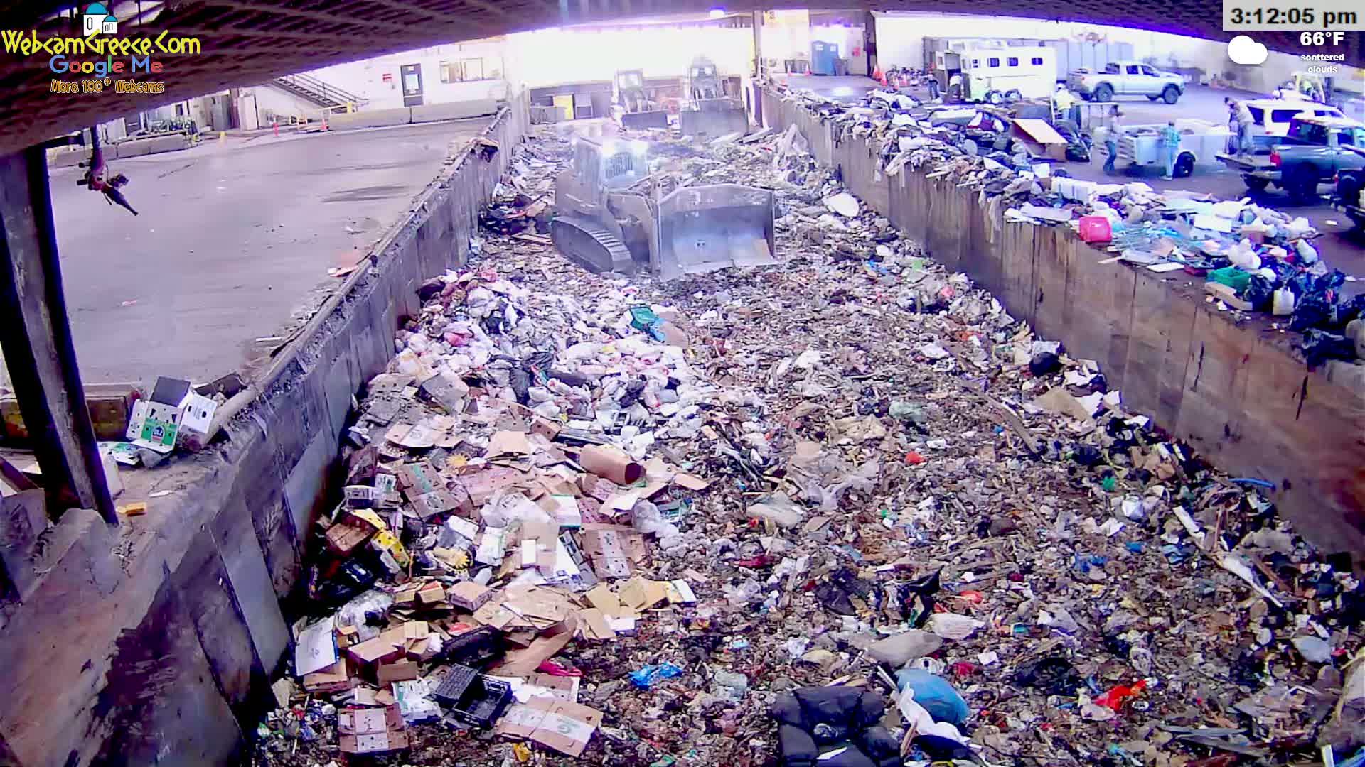 A large pile of trash and debris is being compacted by a machine in an outdoor area under scattered clouds, with a temperature of 66°F.