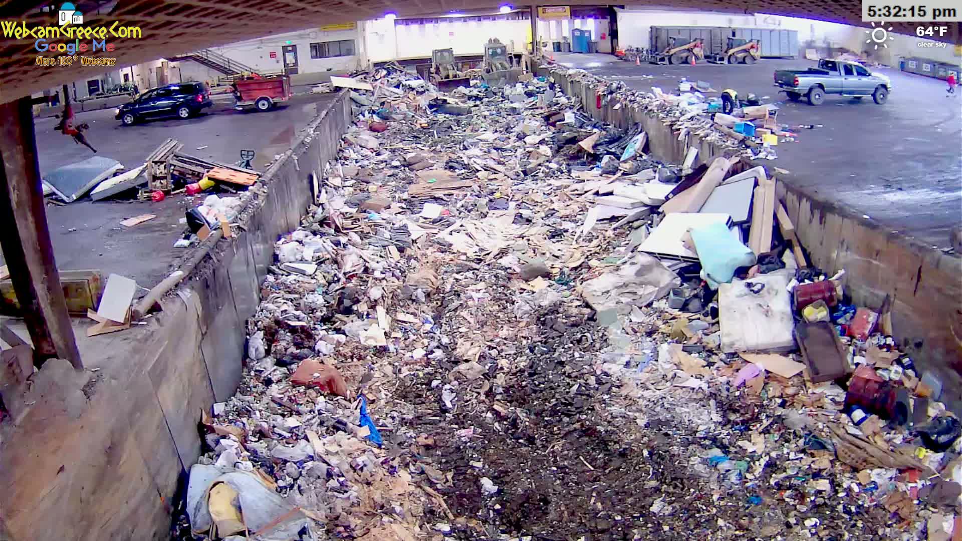 A large pile of refuse is seen in an outdoor, covered area under a clear sky, with vehicles and machinery visible in the background.