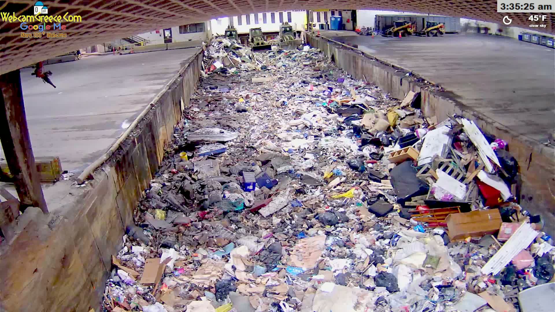A large amount of trash and debris fills a long, narrow, outdoor receptacle under a clear sky.
