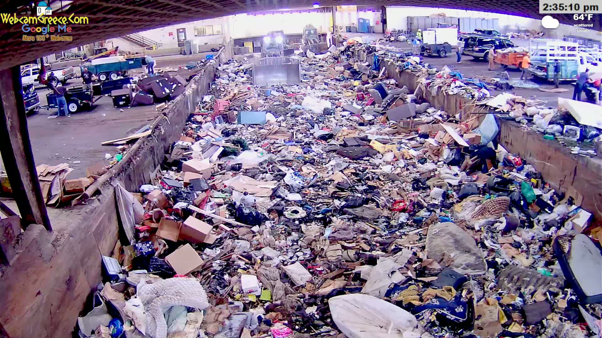 A large amount of trash and debris fills a waste disposal area outdoors under scattered clouds with a temperature of 64°F.