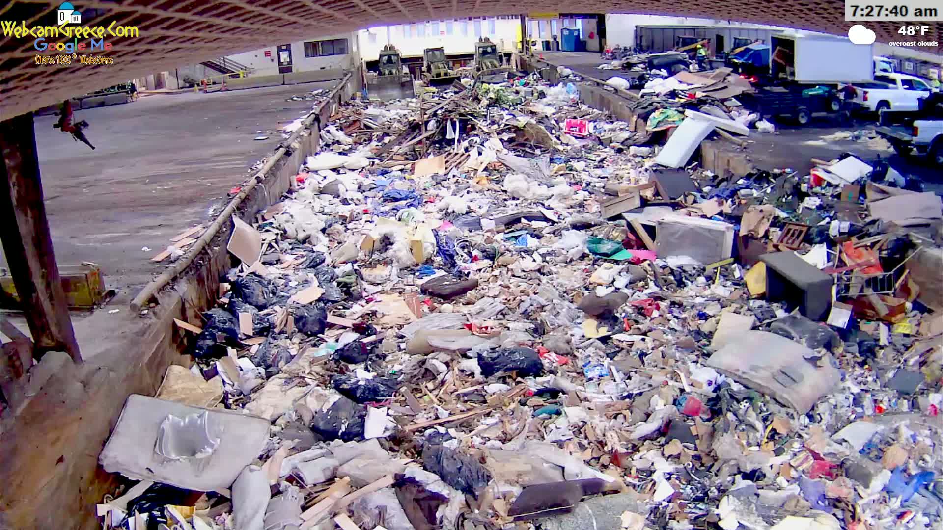 A large pile of garbage sits in an outdoor area under overcast skies, with several pieces of heavy machinery visible in the background.