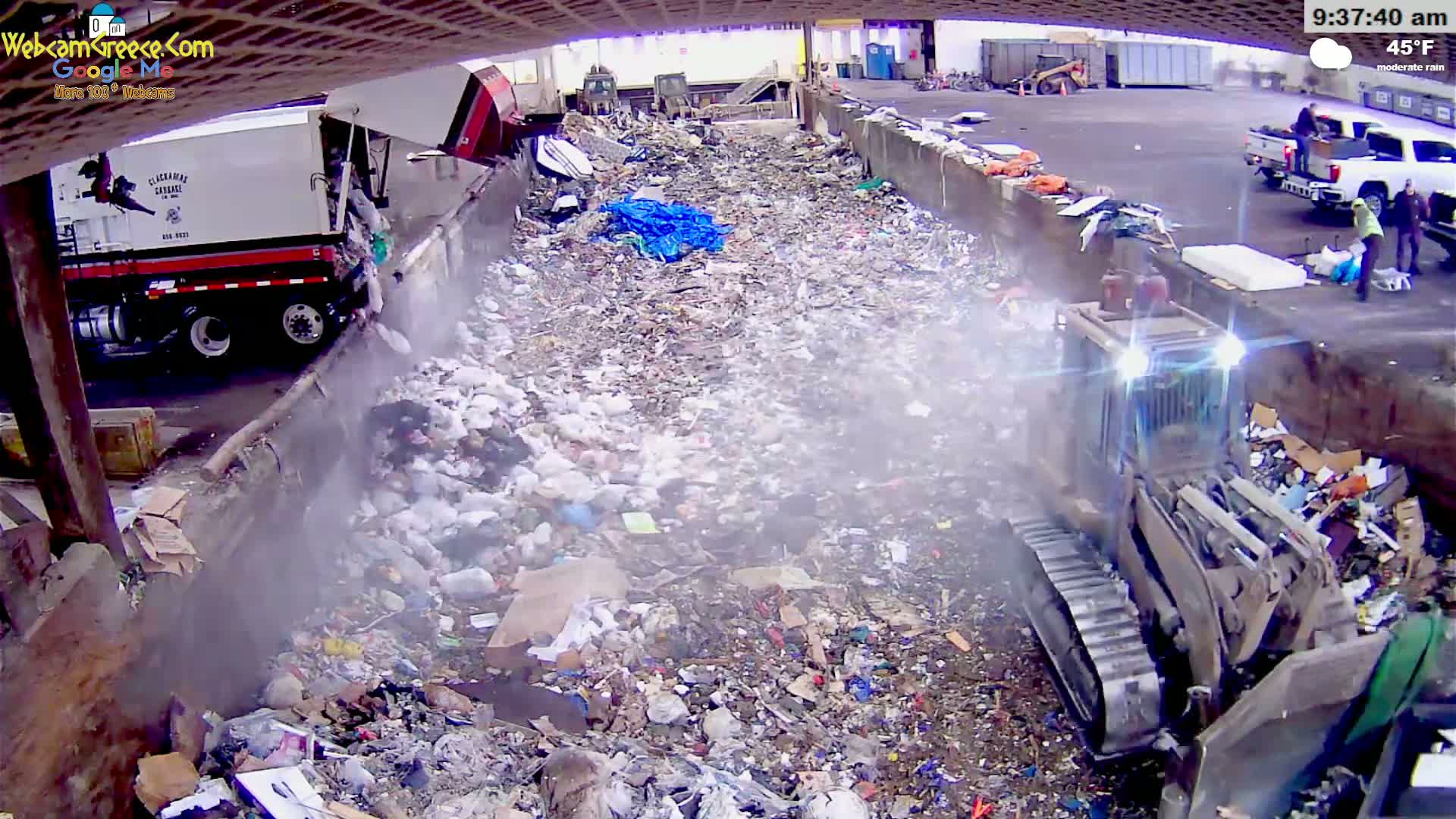 A bulldozer and garbage truck work amidst a large pile of trash in moderate rain.