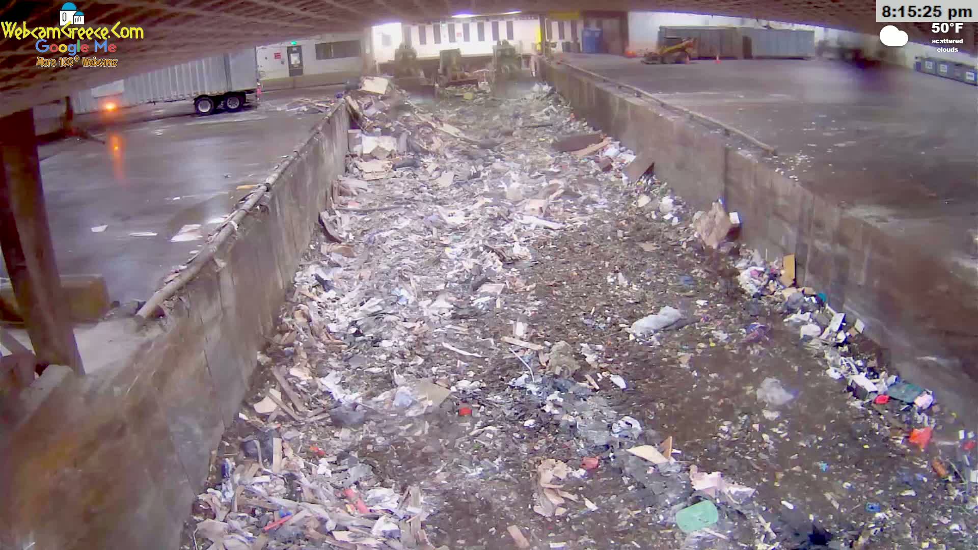 A large amount of trash and debris fills a long, narrow, concrete-walled trough under a partially covered outdoor area with scattered clouds and a temperature of 50°F.