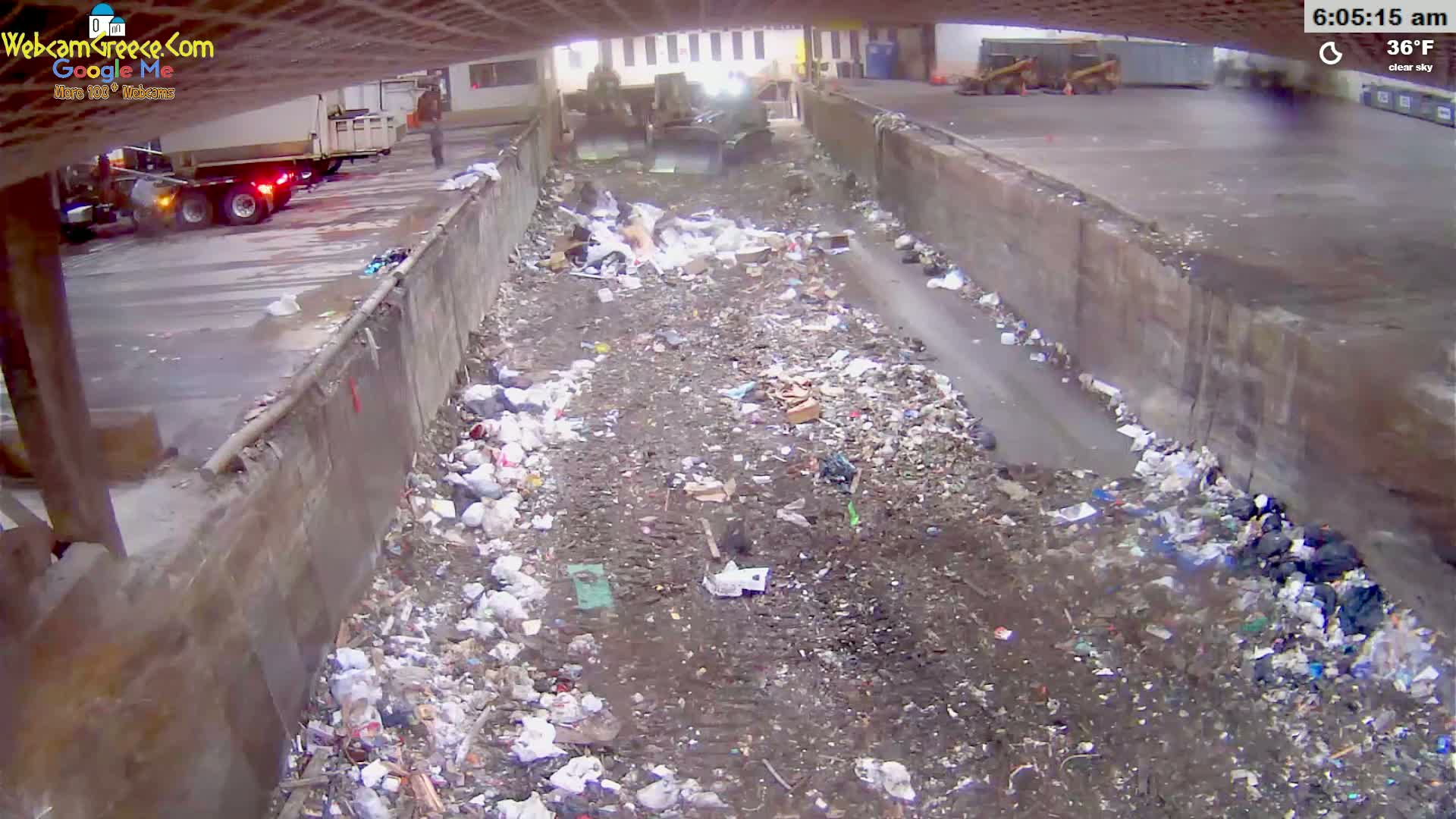 A large amount of garbage and refuse is piled in a concrete-walled channel, with trucks visible in the background under a clear sky.