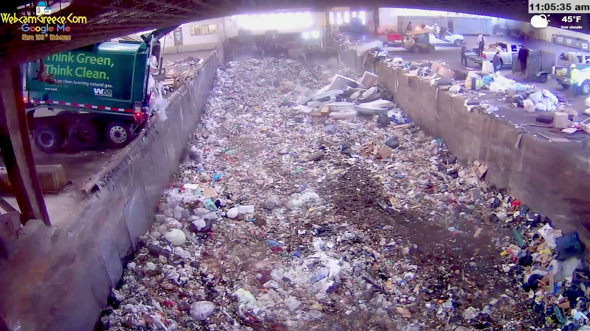 A large pile of trash fills a trench between two concrete walls on a partly cloudy 45°F day, with a green garbage truck partially visible to the left.