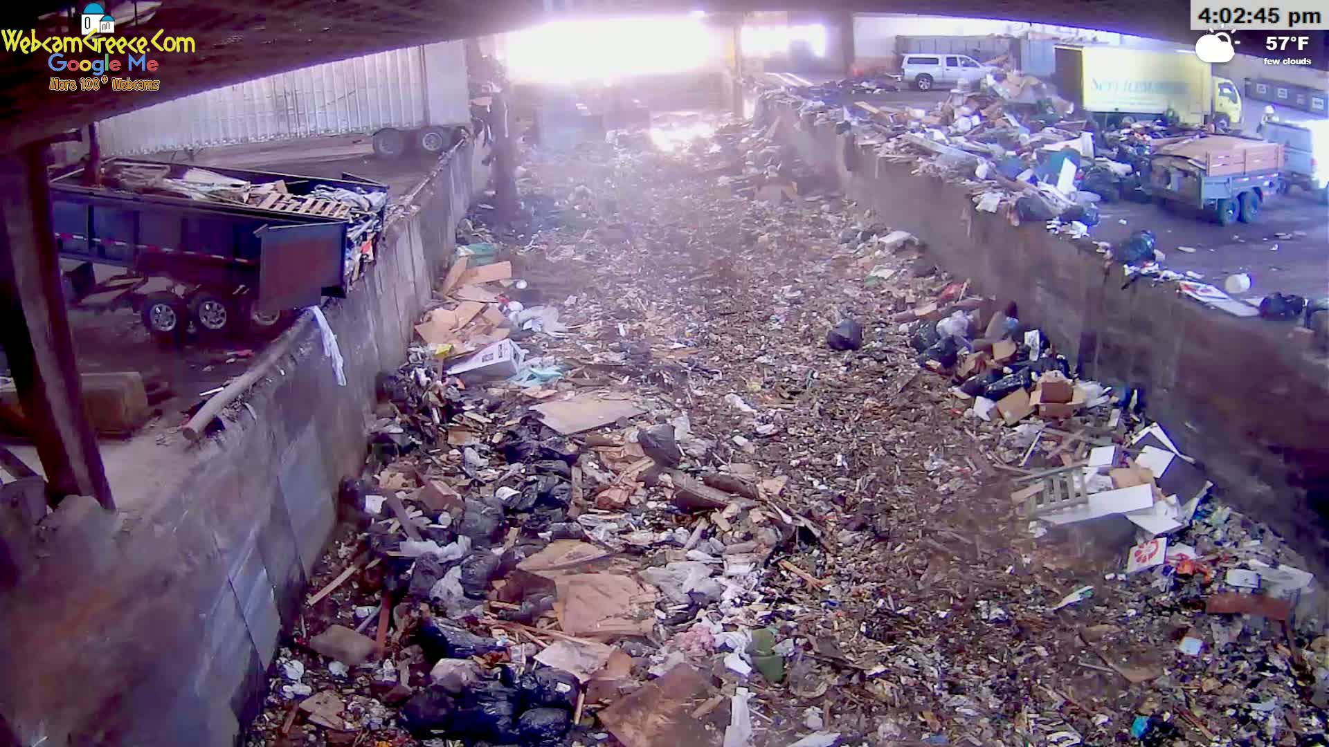 A large pile of trash and debris fills a long, narrow area between two concrete walls under a partly sunny sky.