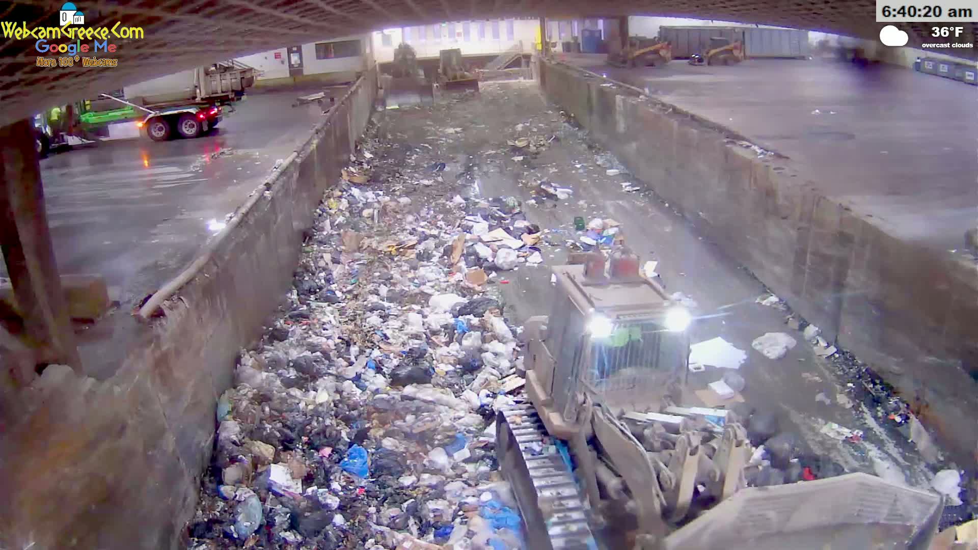 A bulldozer moves through a large pile of garbage in a concrete channel under overcast skies, with a truck visible in the background.