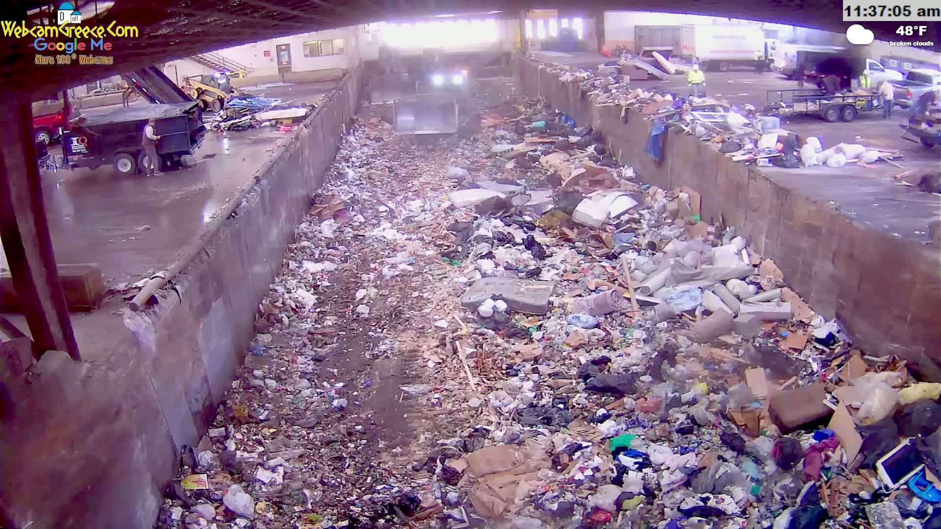 A large amount of trash is piled in a concrete-walled trough under partly cloudy skies, with a piece of machinery visible in the distance.
