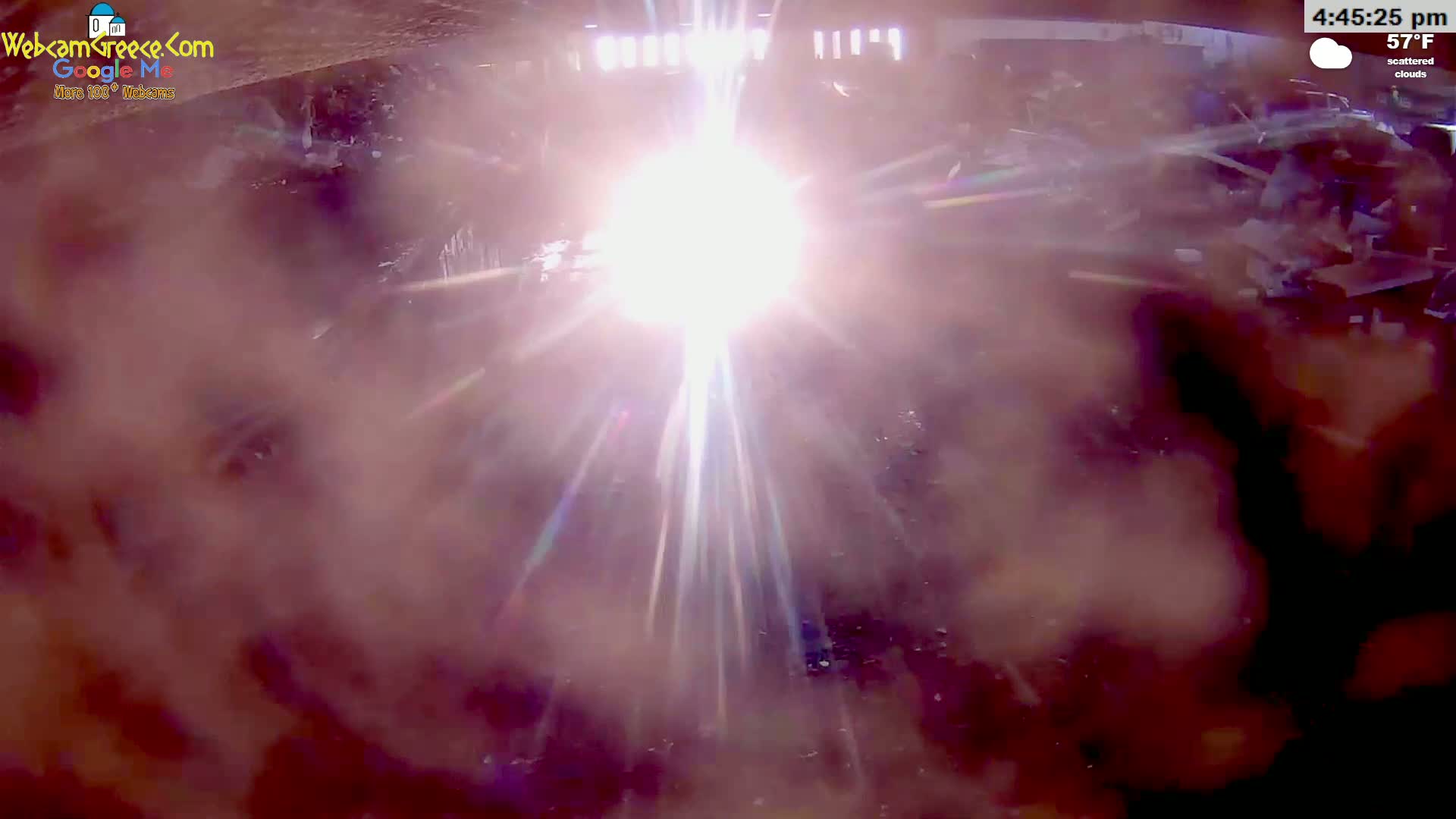 A bright, hazy sun shines down on a dark, cluttered outdoor area under scattered clouds.