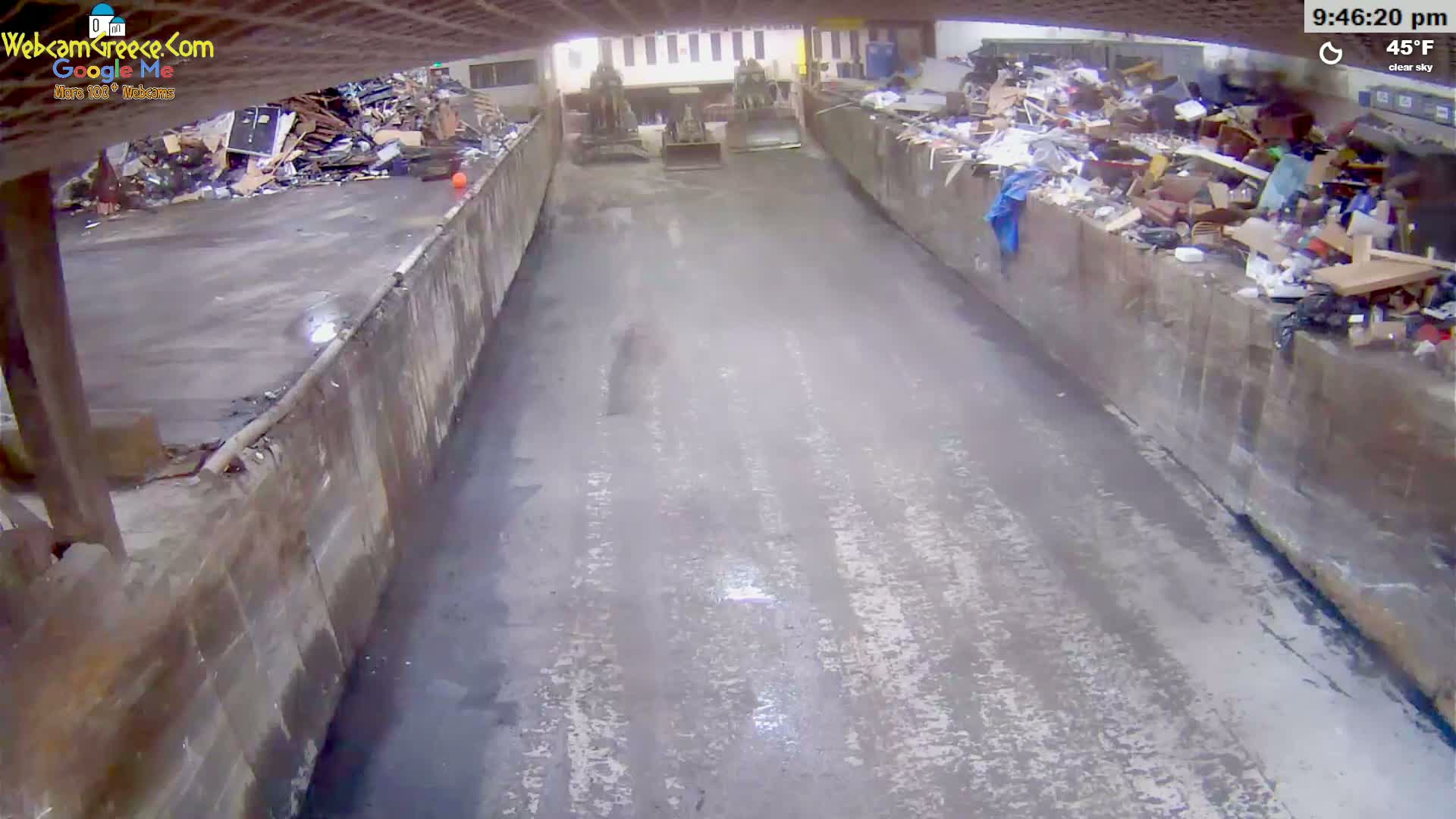 A view down a narrow, paved alleyway between two high concrete walls filled with piles of refuse, with two small pieces of construction equipment visible in the distance under a clear sky at 45°F.