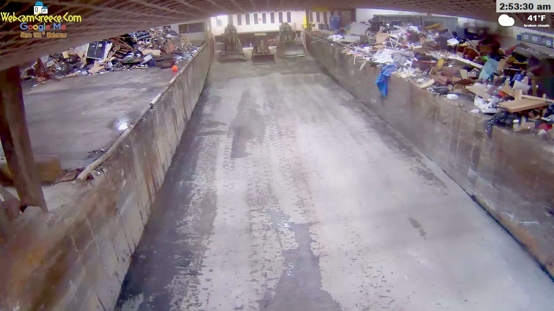 A wet concrete roadway is flanked by large containers filled with refuse, and two pieces of heavy machinery are parked in the middle of the road under broken clouds at 41°F.