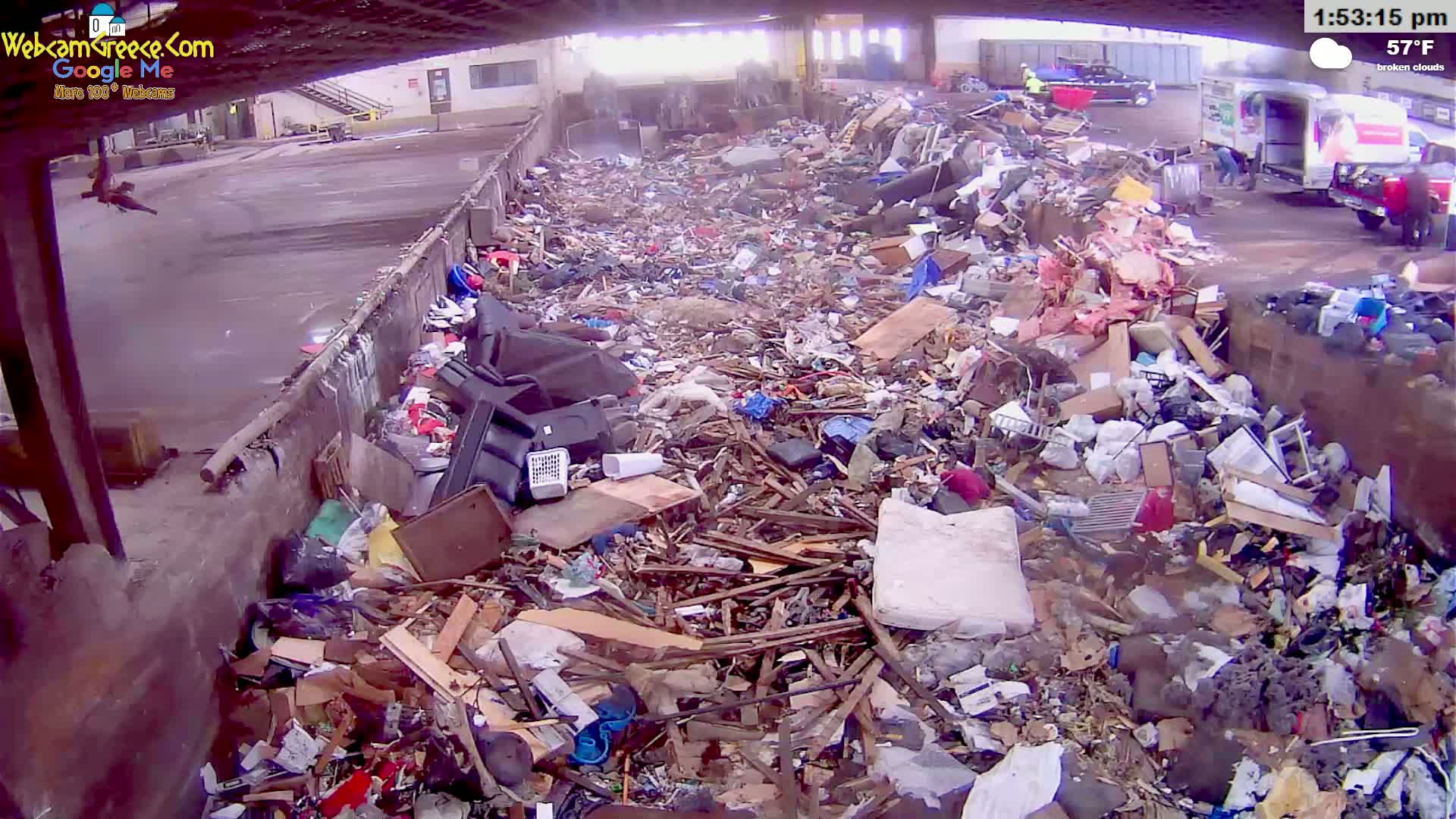 A large pile of assorted debris, including wood, plastic, and other refuse, fills a large outdoor container under broken clouds at 57°F.