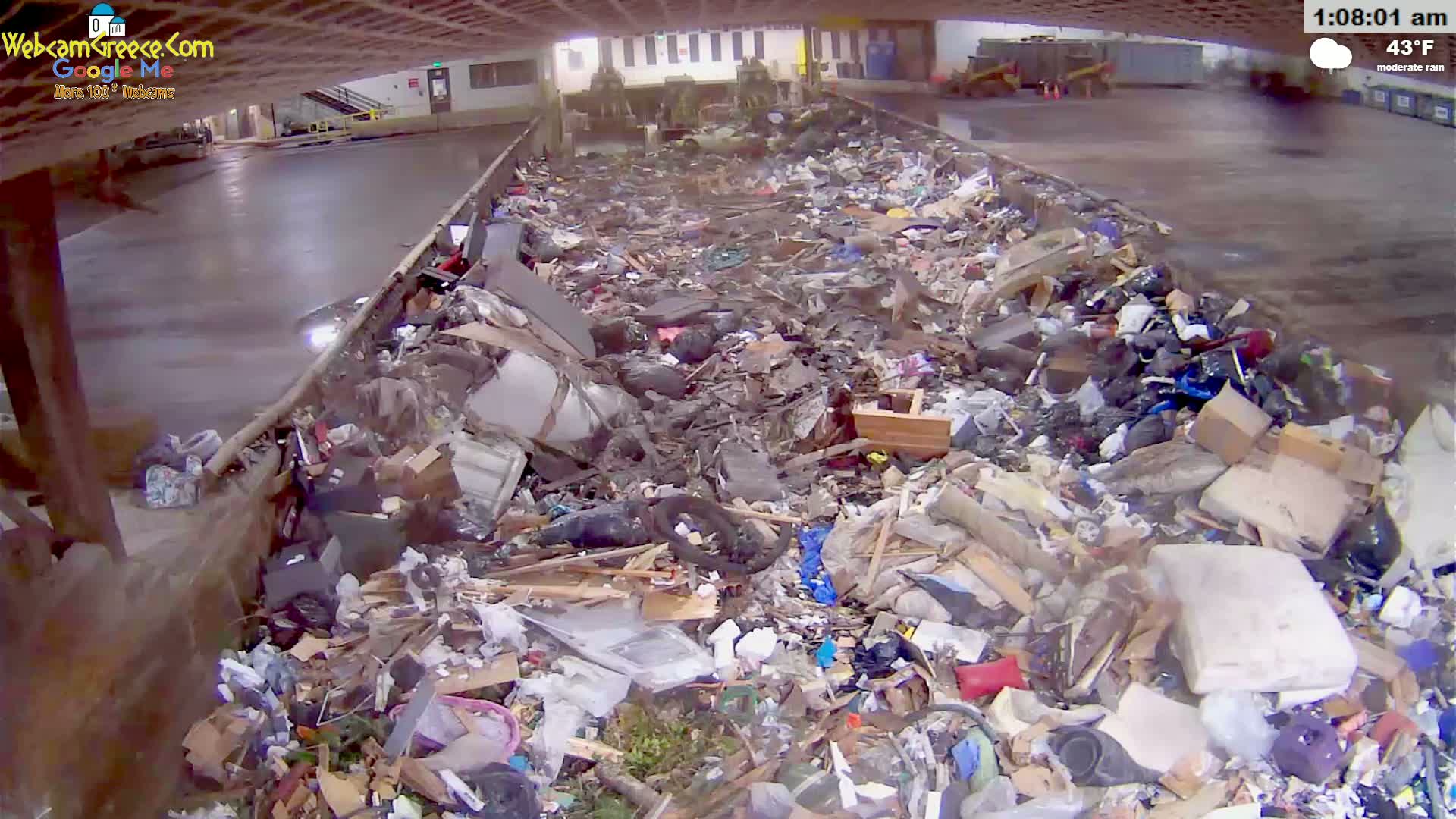 A large pile of trash and debris sits inside a covered structure during moderate rain.