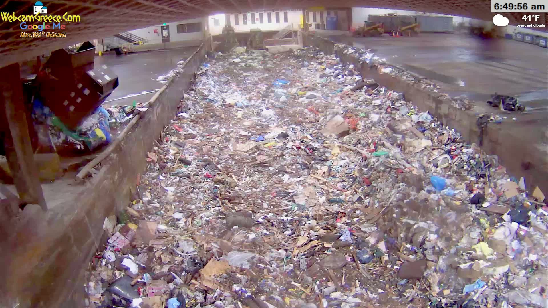 A large amount of garbage fills a long, open-topped trough under overcast skies.
