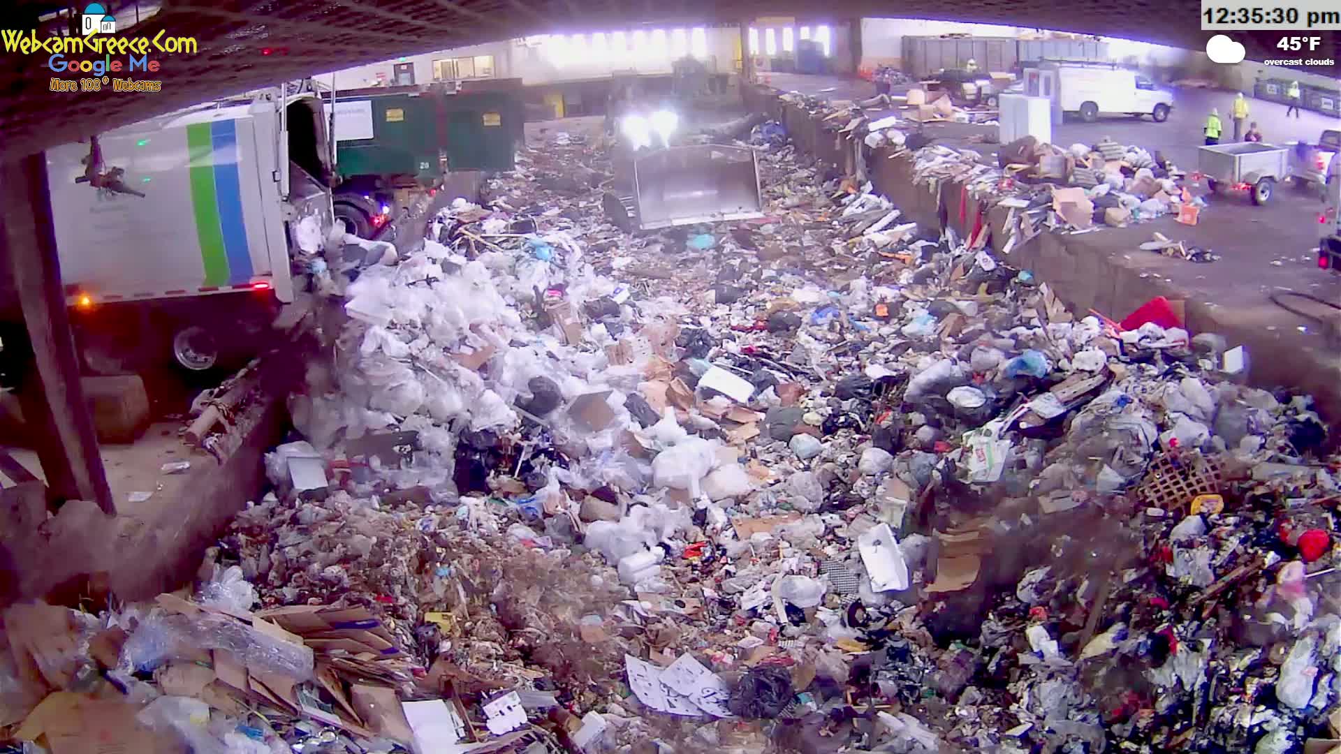 A garbage truck dumps trash into a large pile of refuse on an overcast day, with the temperature at 45°F.