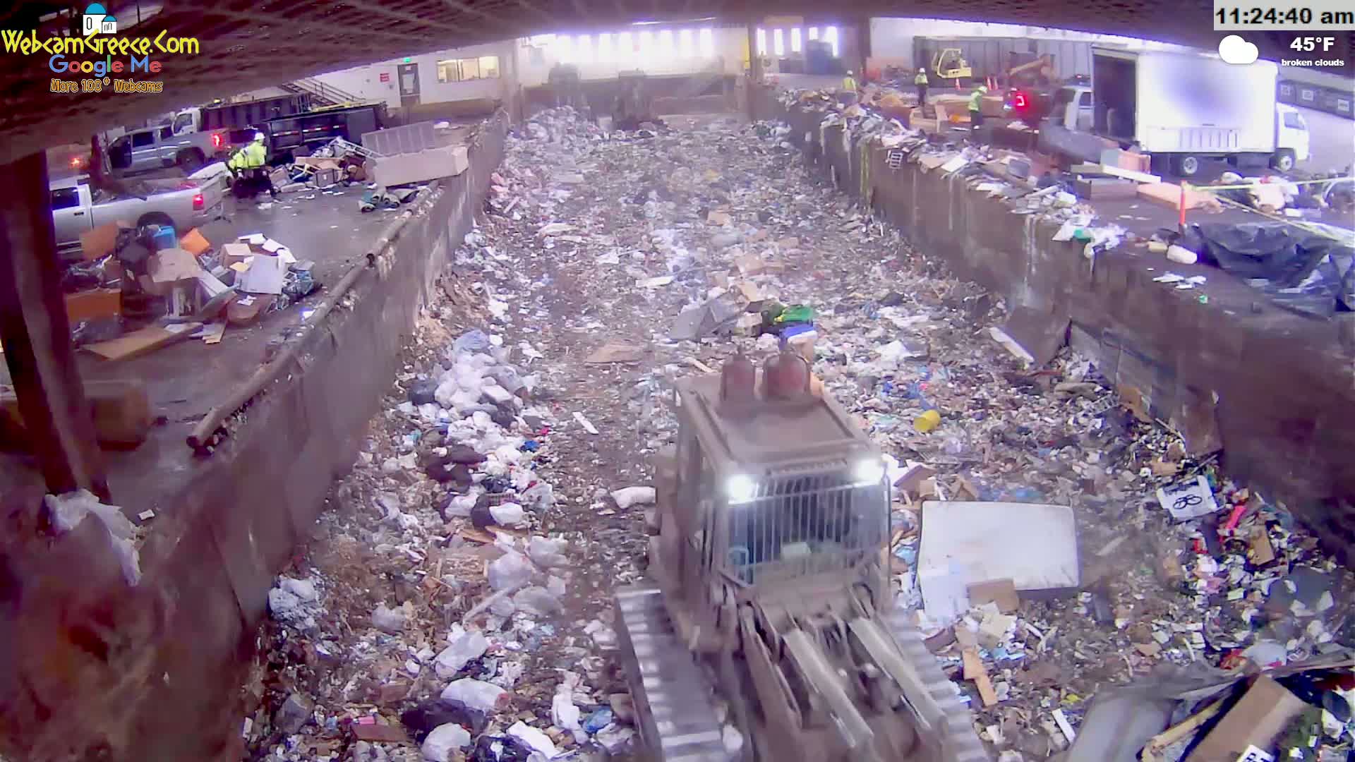 A bulldozer moves through a large pile of trash in an outdoor landfill under broken clouds.