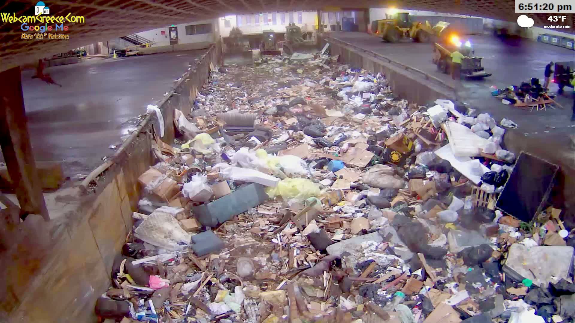 A large pile of garbage sits inside a waste disposal facility under moderate rain, with yellow machinery visible in the background.