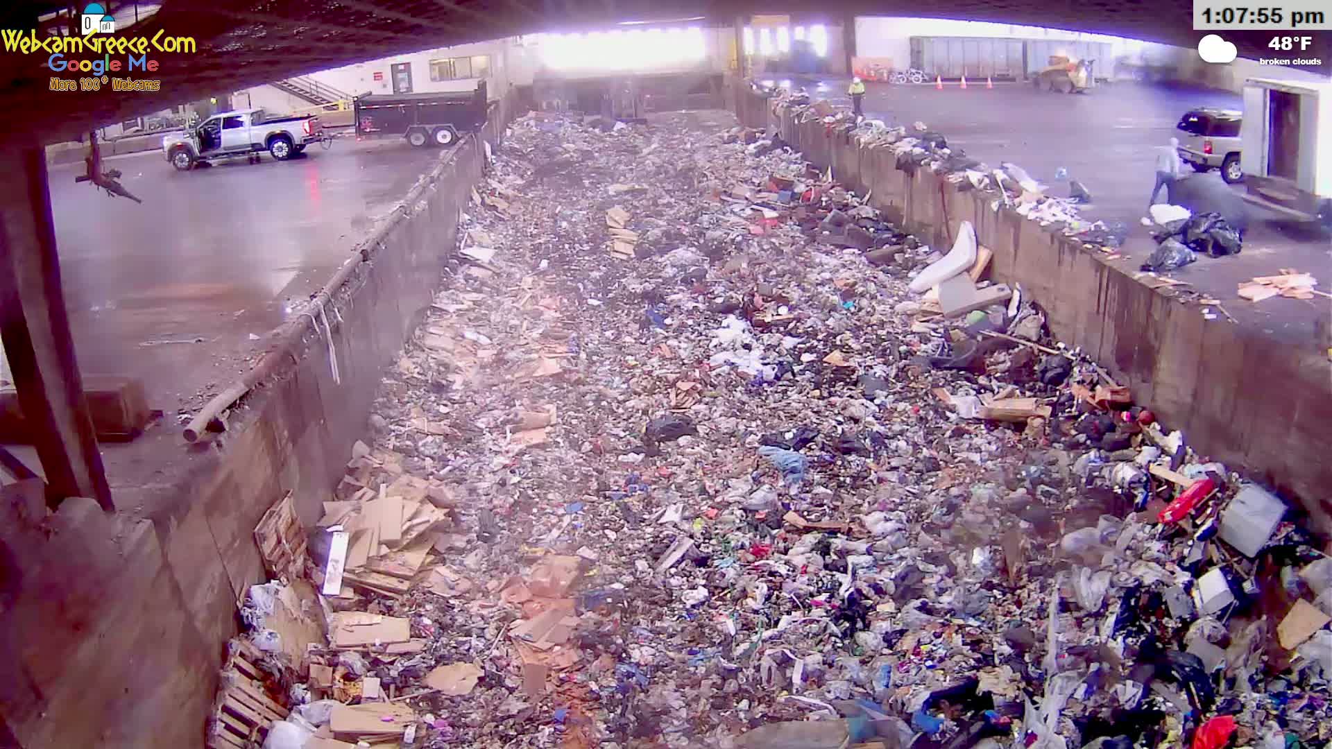 A large pile of trash sits between two concrete walls under broken clouds, with a truck and a person visible in the background.