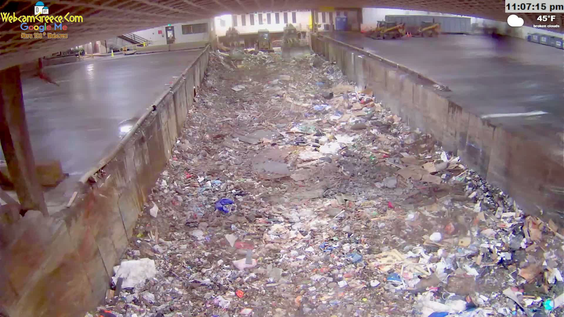 A large pile of trash sits in a concrete-walled area under a partially visible overpass on a day with broken clouds and a temperature of 45°F.