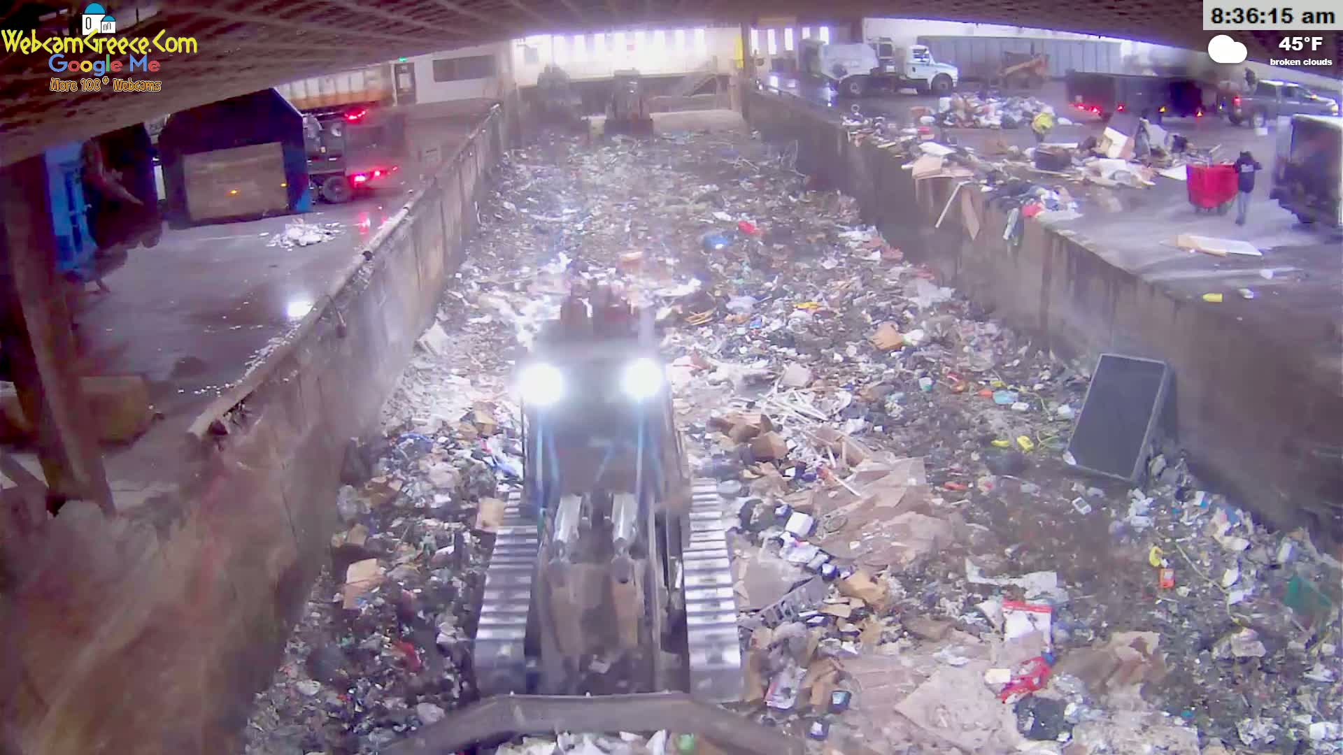 A small bulldozer moves through a large pile of trash in an outdoor facility under broken clouds at 45°F.