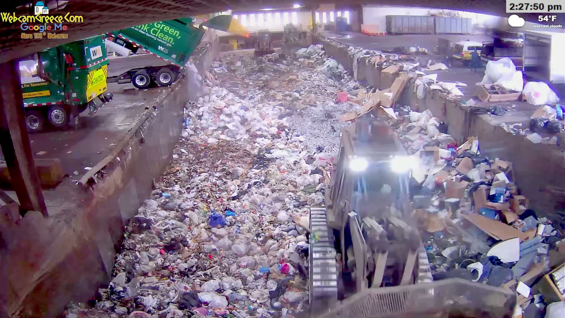 A large pile of garbage is being processed by machinery under overcast skies at 54°F.