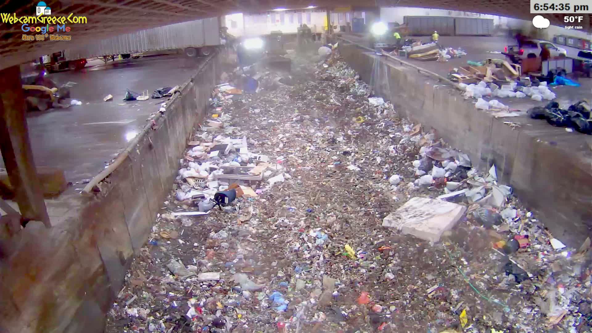 A large pile of trash and debris fills a concrete-walled pit under a slightly overcast sky with light rain.