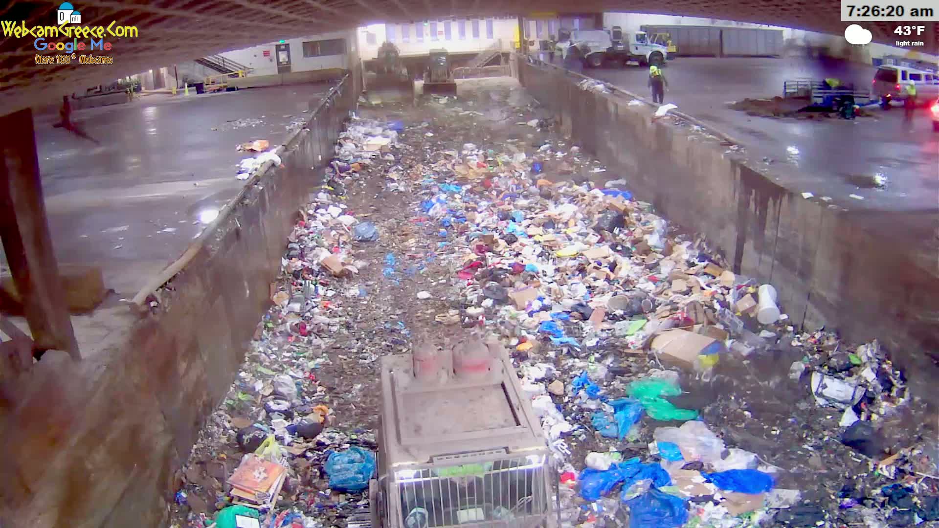 A large pile of trash sits in a concrete trough under a covered area during light rain, with a garbage truck positioned in the center of the pile.