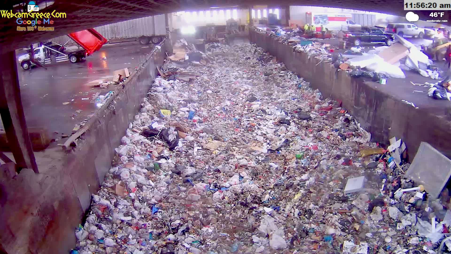 A large pile of garbage sits in a trough under an overpass during moderate rain.
