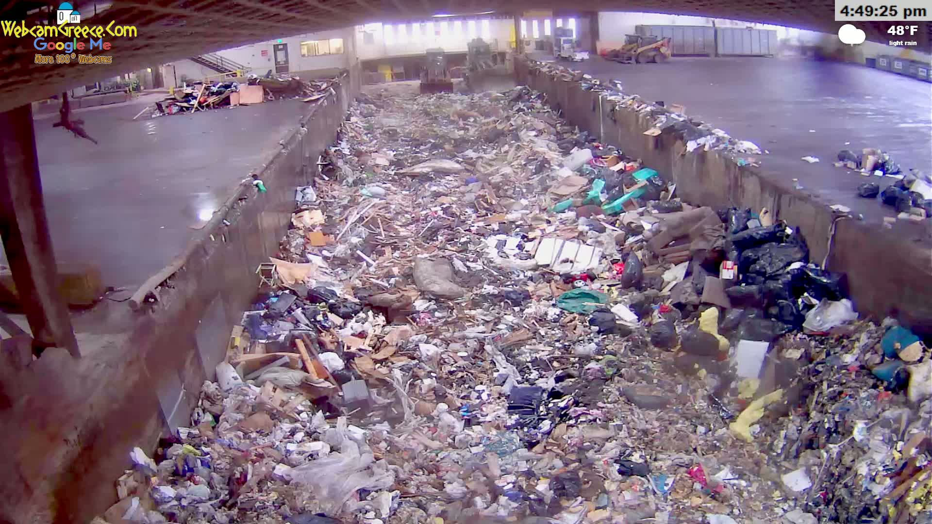 A large pile of trash and debris fills a long, narrow concrete trough under a covered structure during light rain.