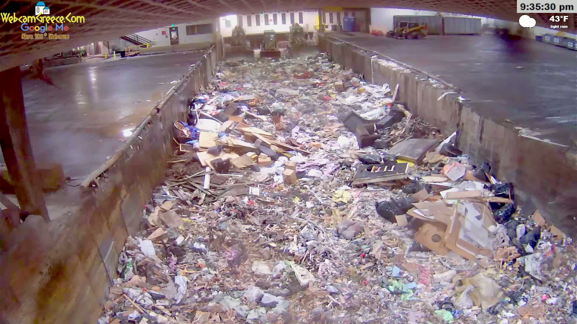 A large pile of trash and debris fills a concrete pit under a partly covered structure during light rain at 43°F.