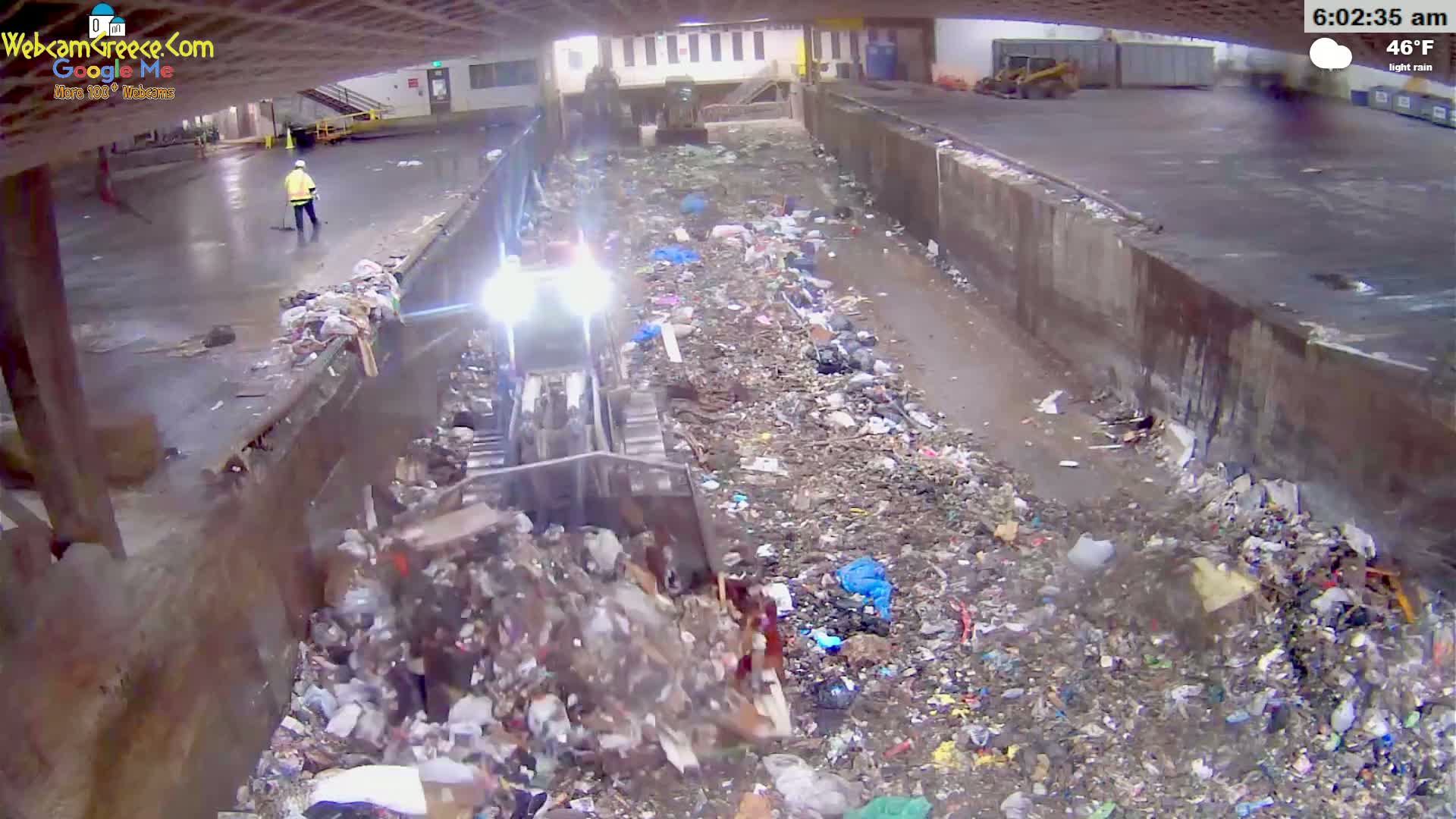 A bulldozer pushes trash in a wet, indoor landfill under light rain at 46°F.