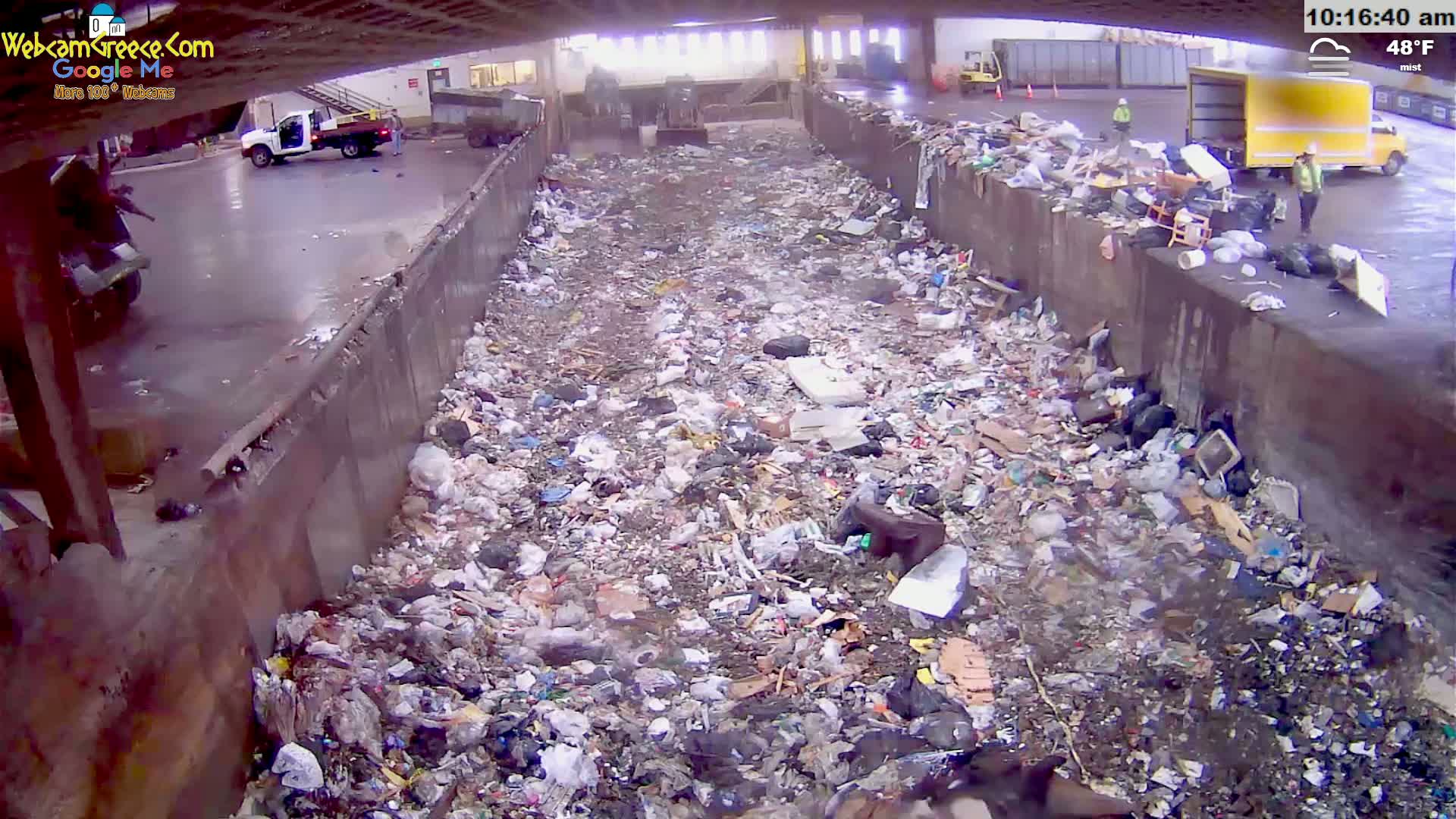 A large pile of trash is seen in a misting, outdoor waste disposal area, with some vehicles and workers visible in the background.