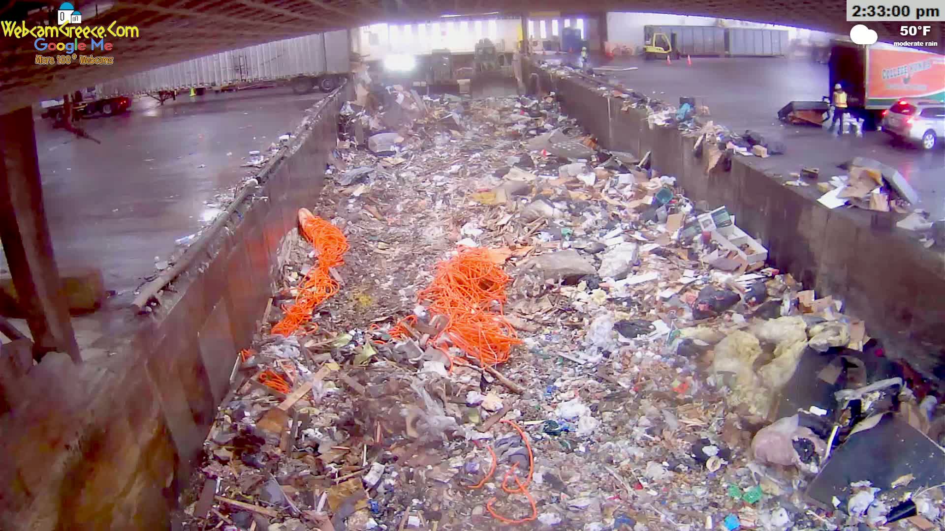 A large pile of refuse and recycling, including bright orange cords, fills a concrete trough outdoors in moderate rain.