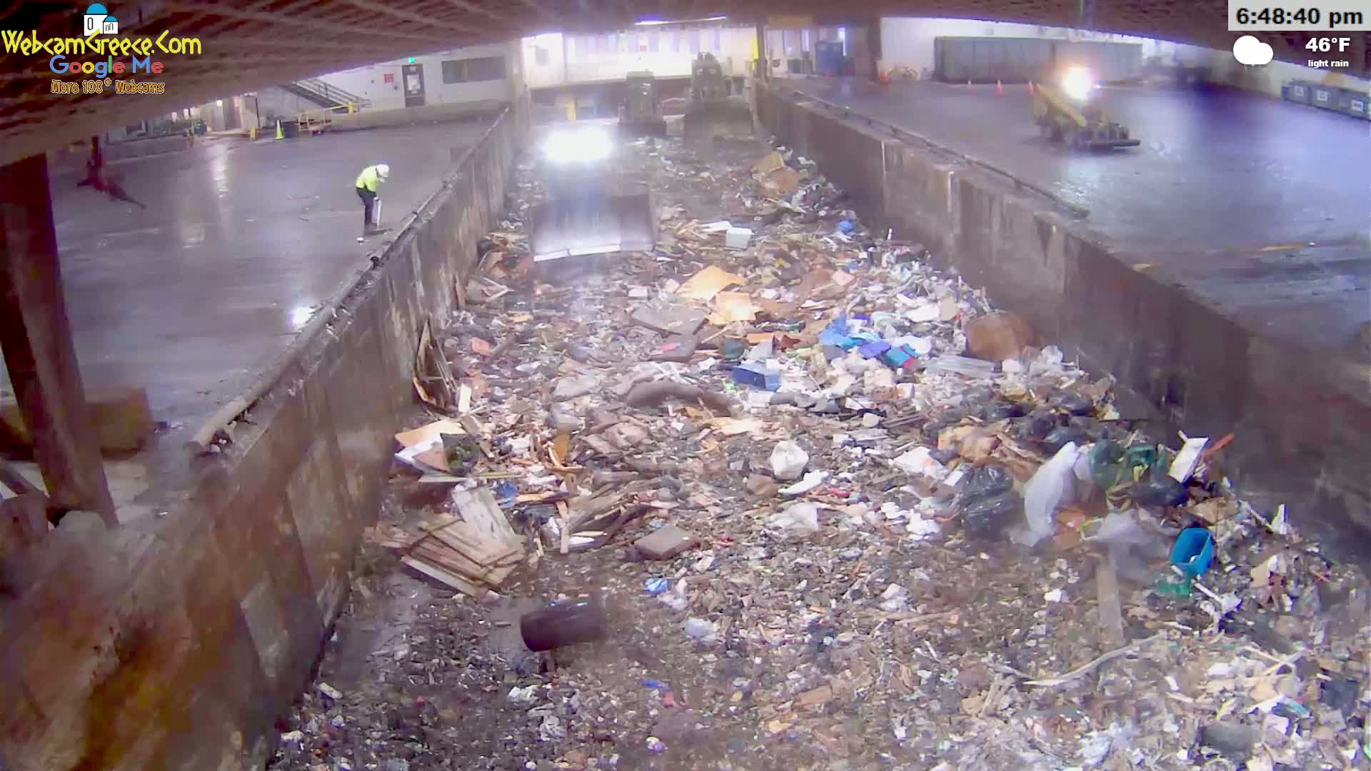 A large pile of garbage is seen in a concrete pit under a covered area during light rain.