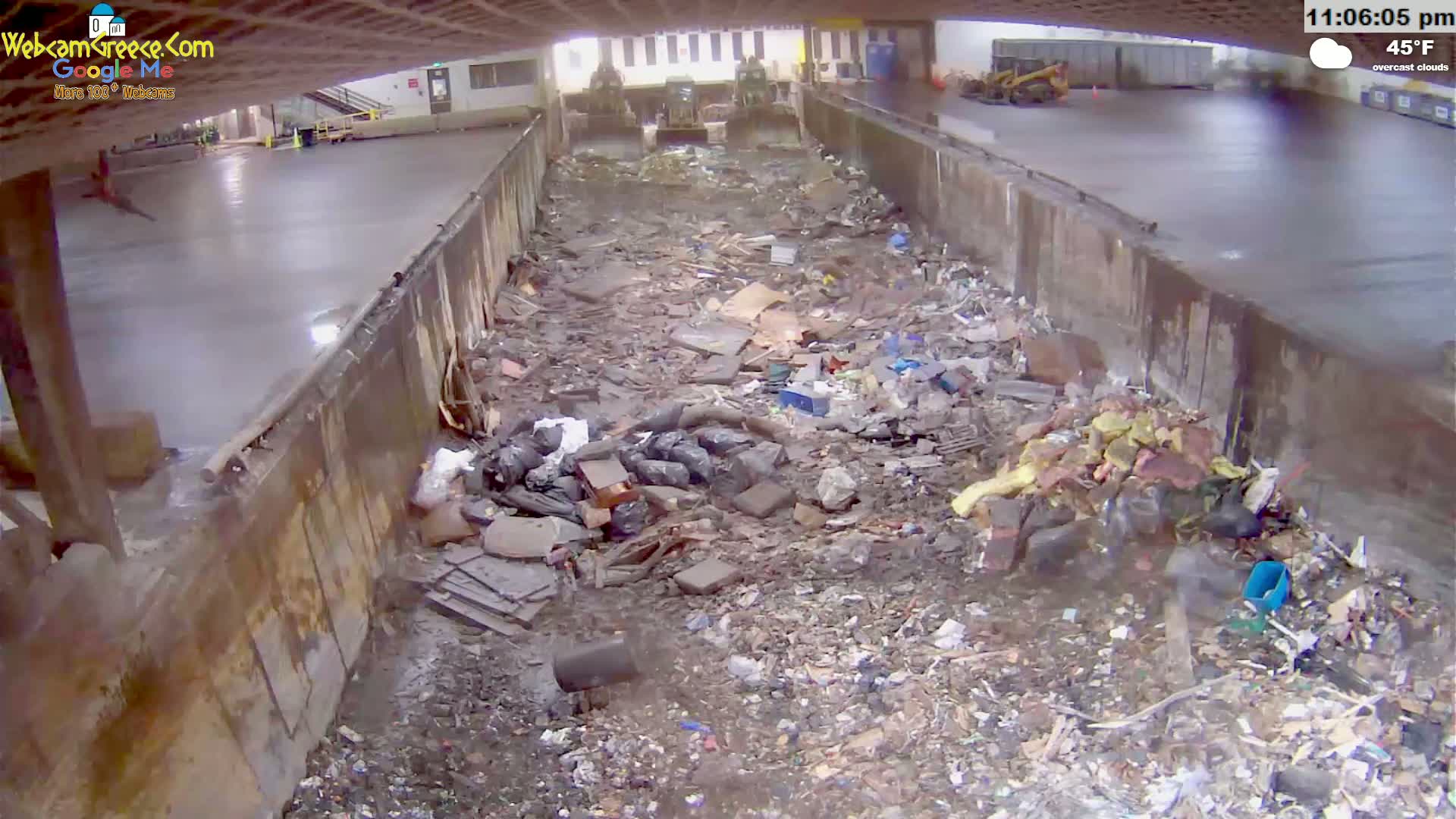 A large pile of trash and debris fills a concrete-lined trough under an overcast sky, with construction equipment visible in the background.