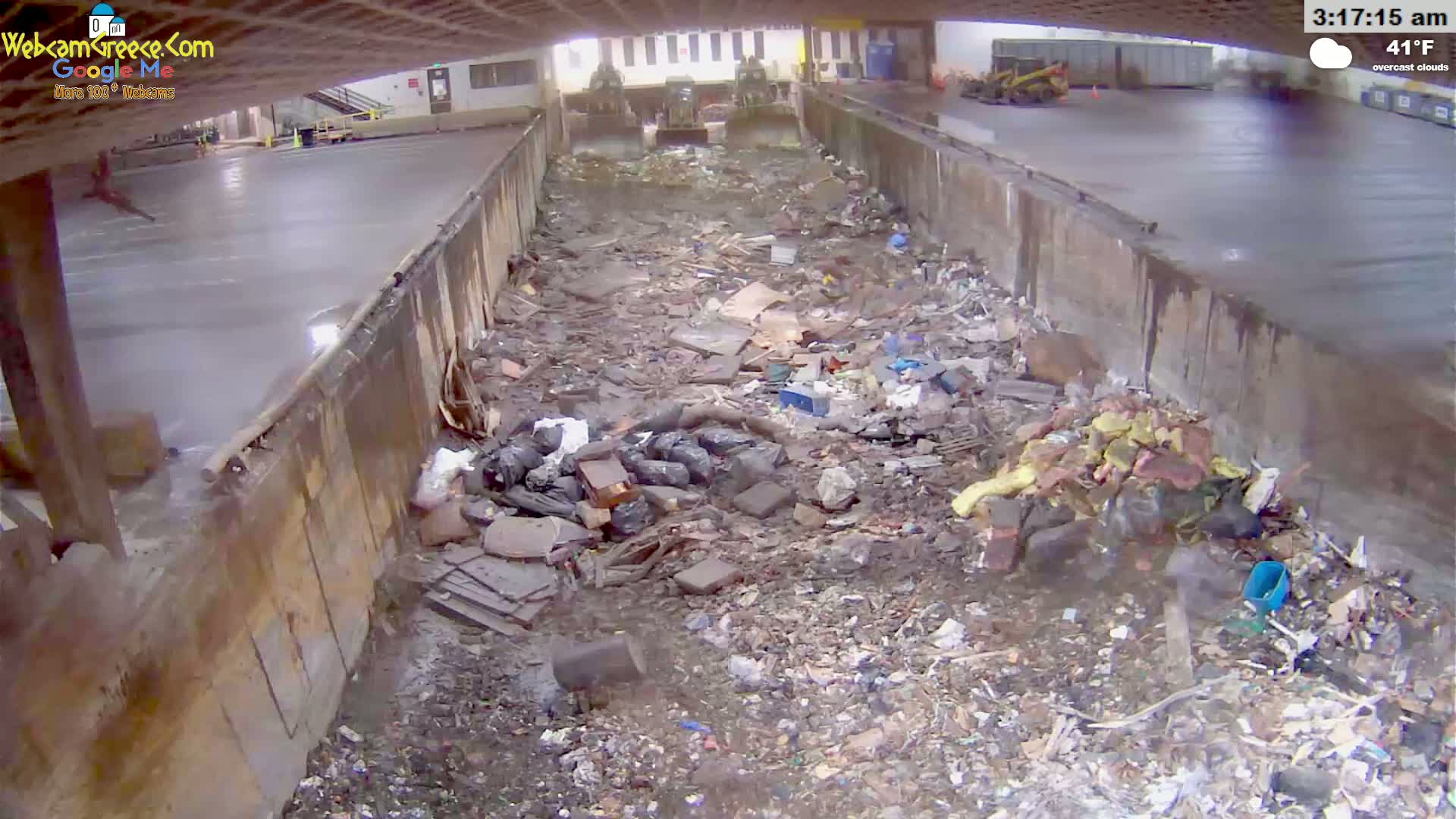 A large amount of trash and debris is piled in a concrete-walled, open-air area under overcast skies, with two pieces of heavy machinery visible in the background.