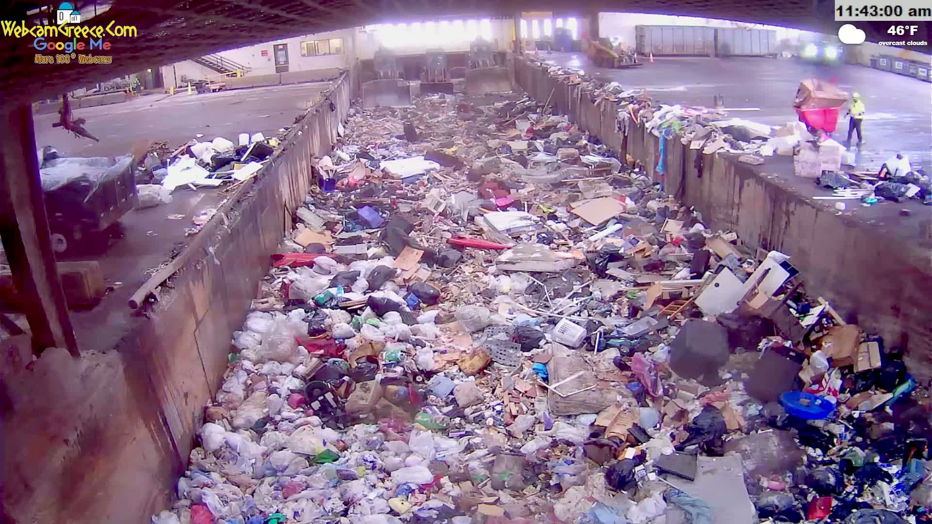 A large pile of refuse sits in an open-air waste receptacle under overcast skies, with a person in bright safety clothing visible in the background.