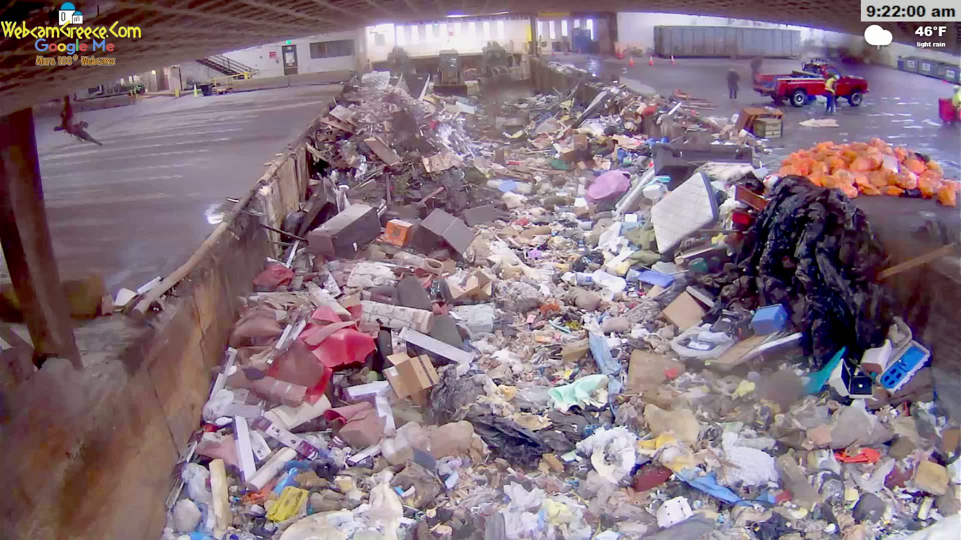 A large dumpster is overflowing with a diverse assortment of trash under light rain.