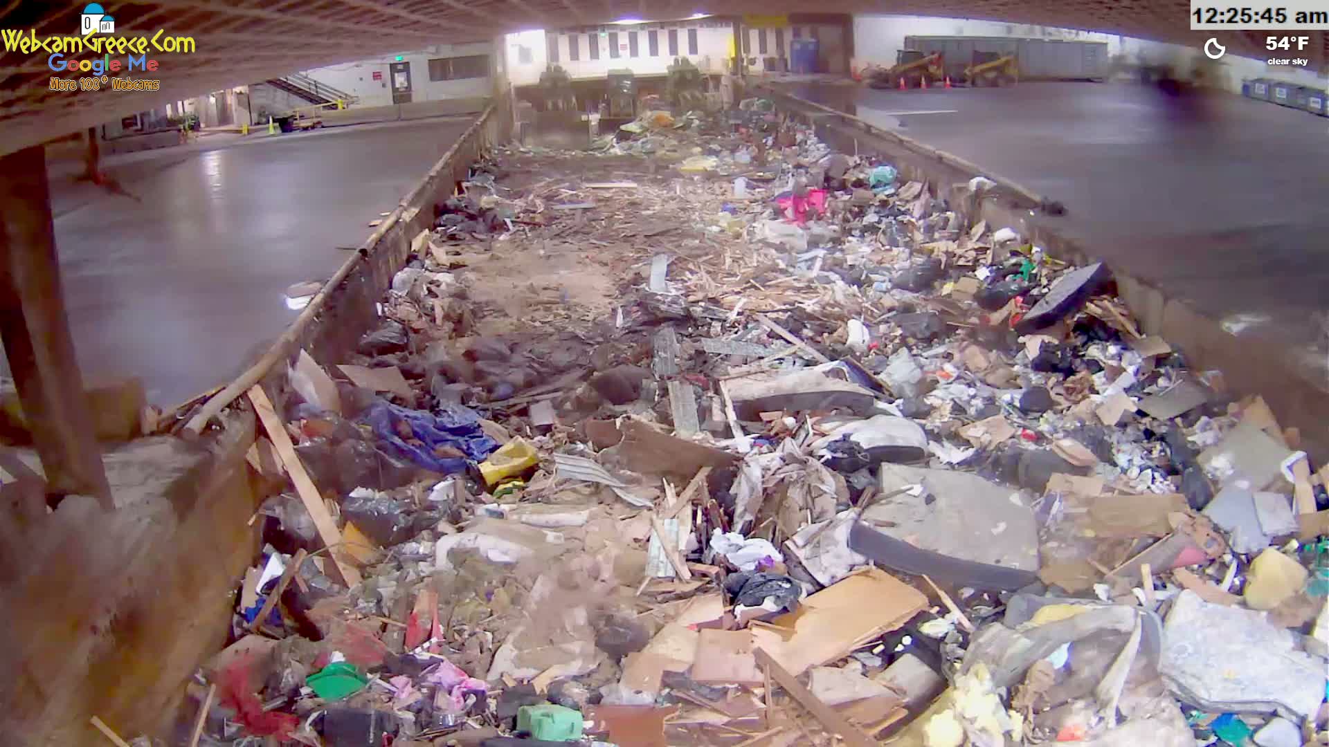 A large pile of trash and debris fills a long, narrow container under a clear sky, with the temperature at 54°F.