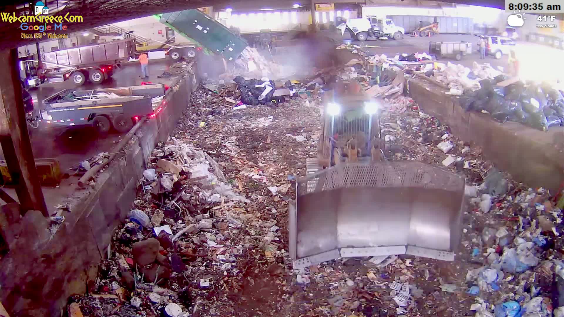 A bulldozer is working in a waste recycling facility on a partly cloudy day, with dump trucks unloading garbage.