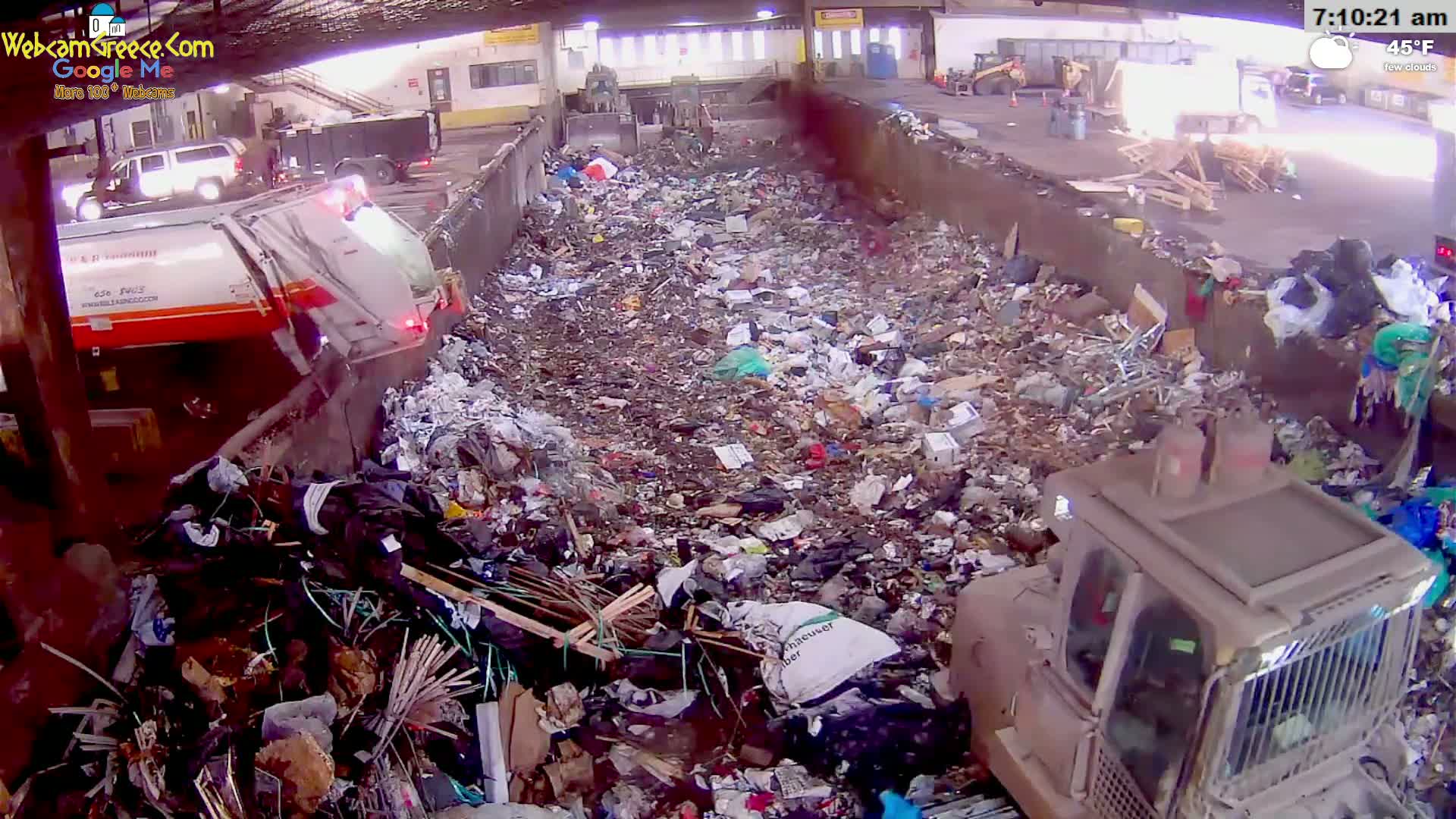 A large pile of garbage is seen inside a waste facility on a cool, partly cloudy morning, with a garbage truck and a bulldozer present.