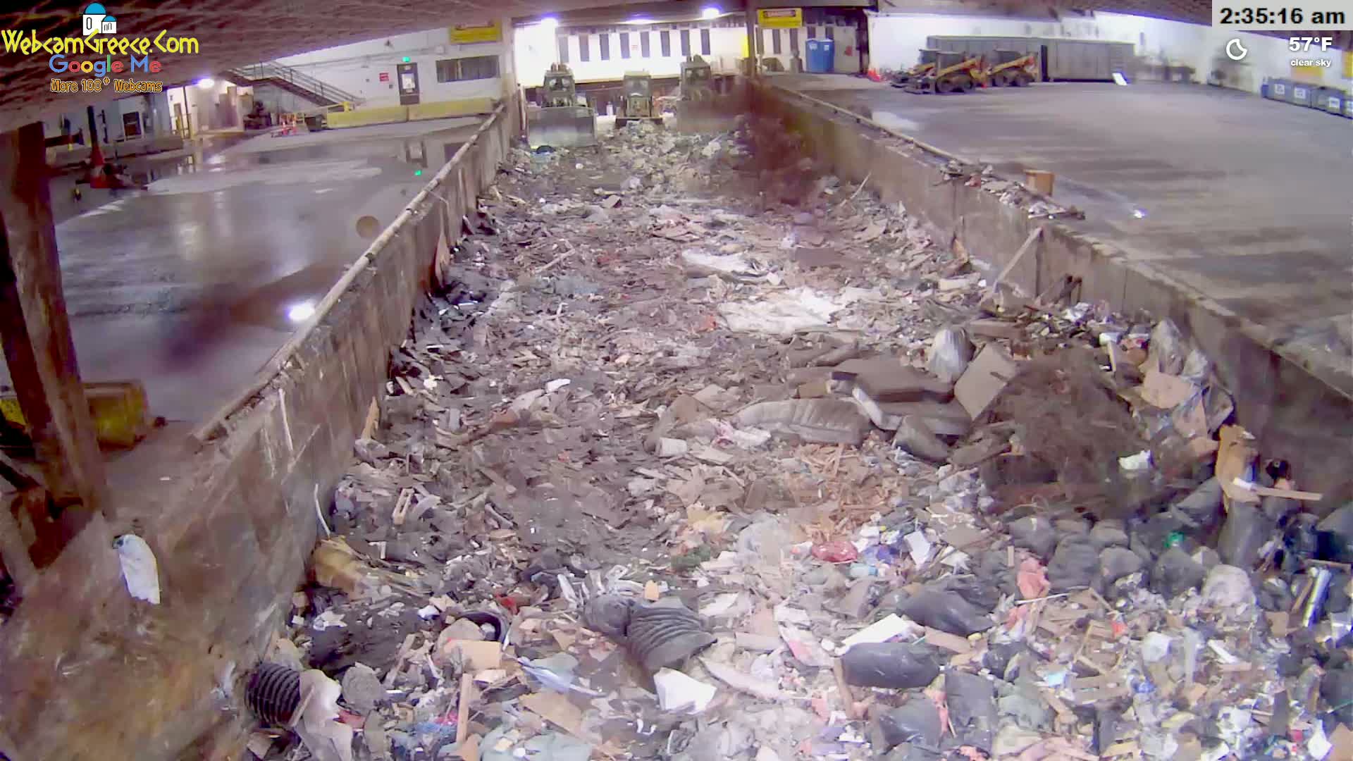 A large pile of mixed waste sits in an indoor concrete trough under a clear sky.