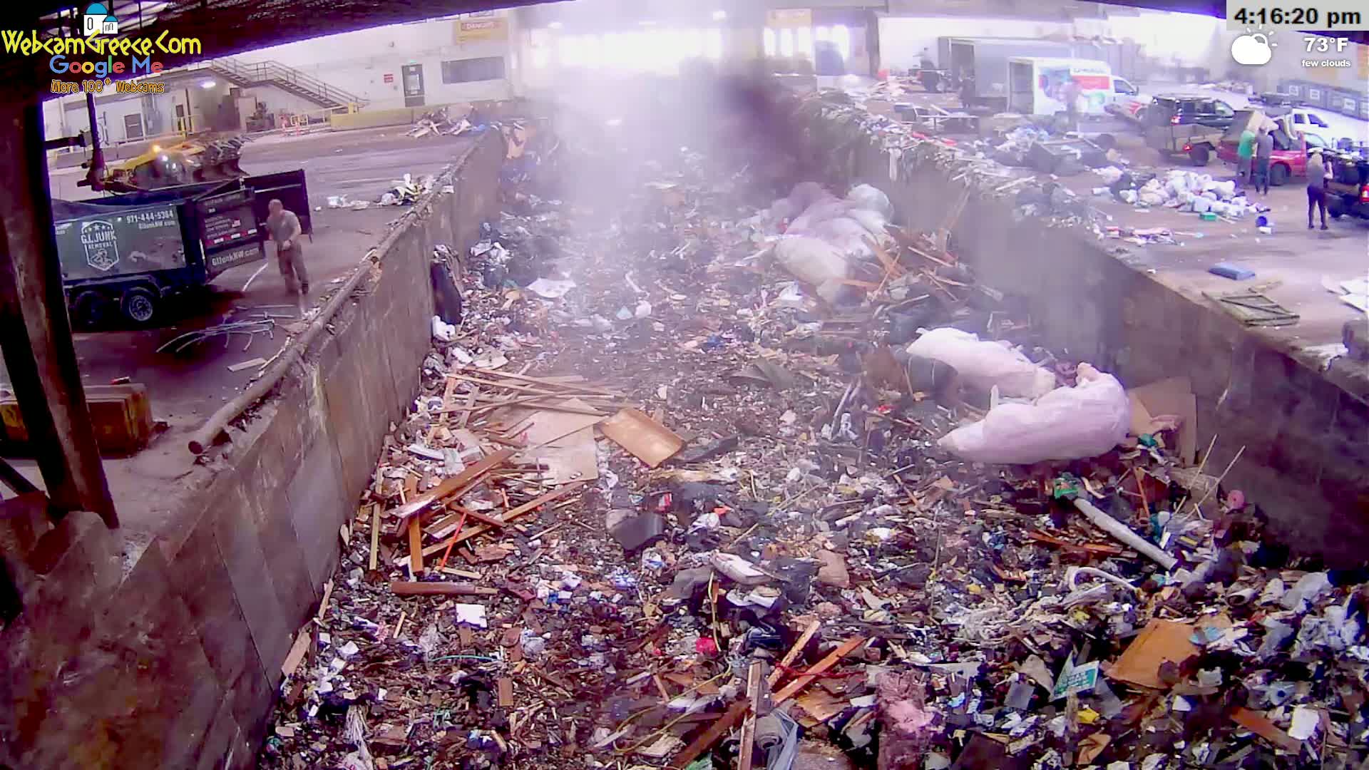 A large dumpster is overflowing with a mixture of wood, debris, and large plastic bags under a partly cloudy sky.