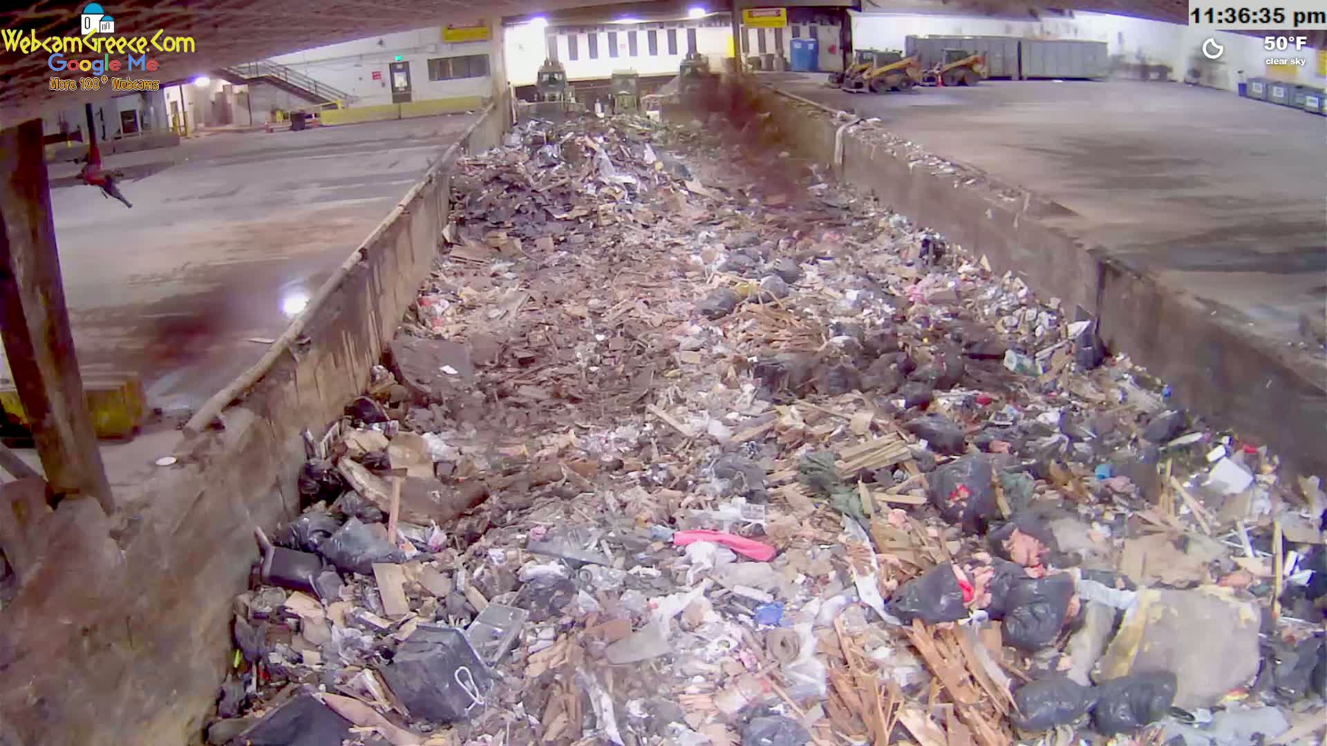 A large pile of garbage fills a long trough-like structure inside a building under a clear sky, with the temperature at 50°F.